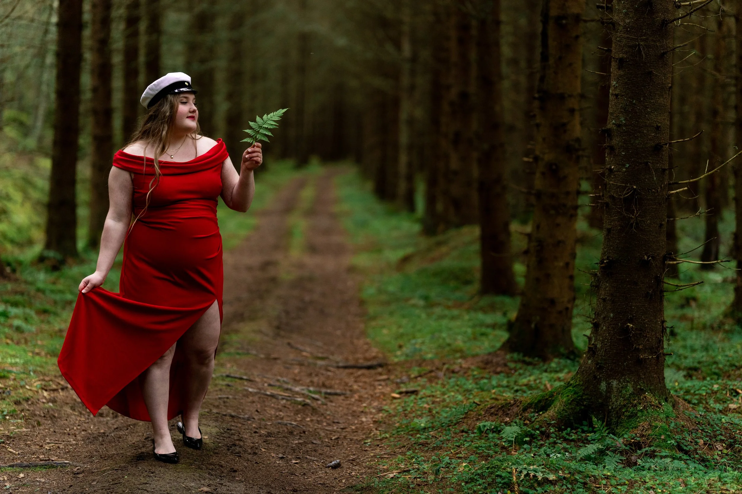 A woman in a red dress and a white hat holding a fern walking in a forest with tall trees.