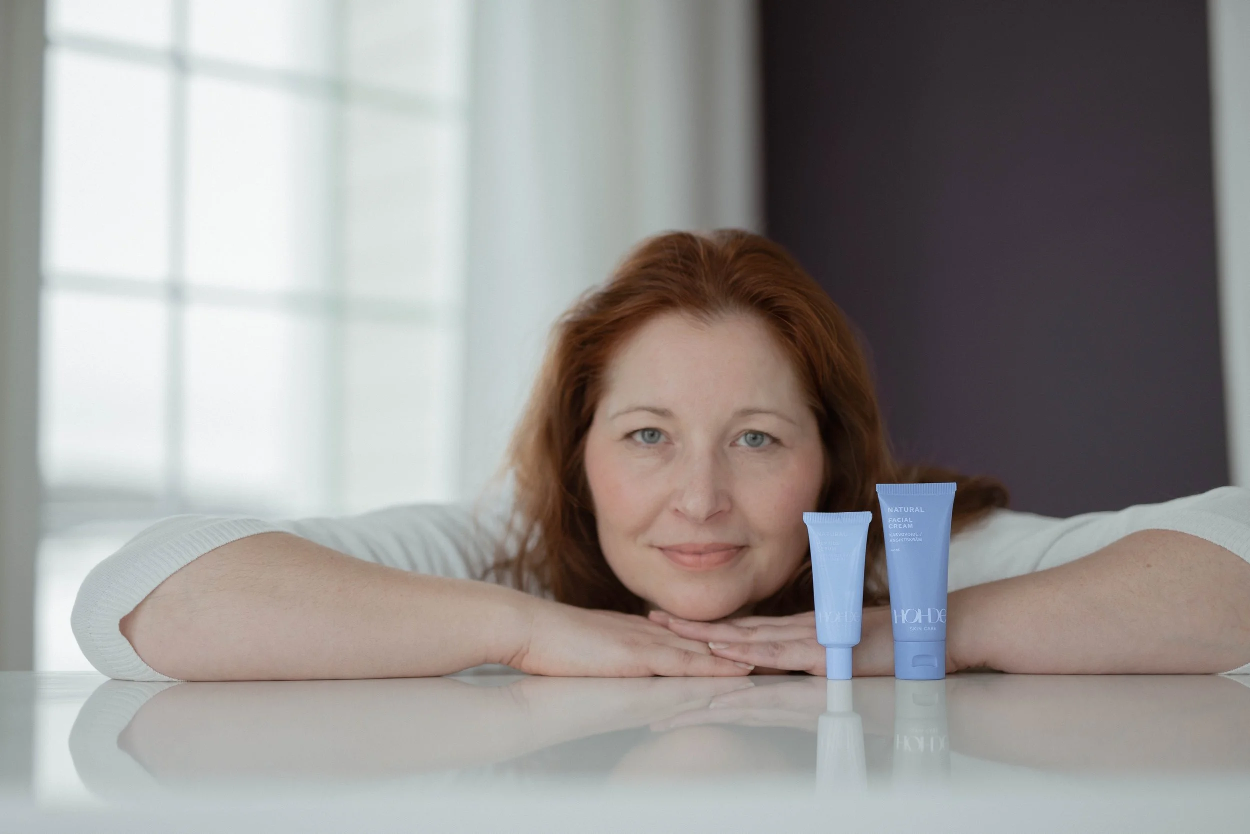 A woman with red hair resting her chin on her hands on a table, with two blue skincare tubes in front of her, smiling gently.