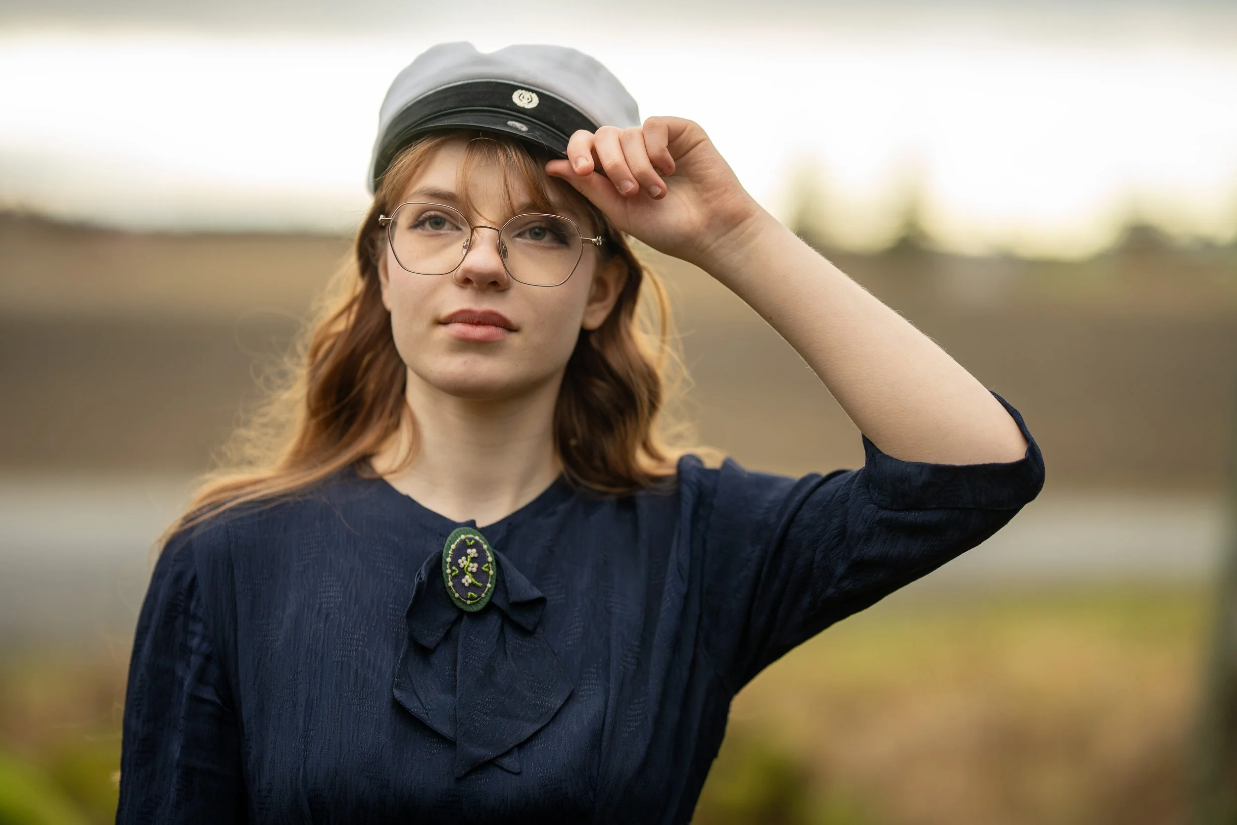 A young woman with red hair and glasses wearing a gray hat and a dark blue blouse with a decorative brooch, standing outdoors and adjusting her hat.