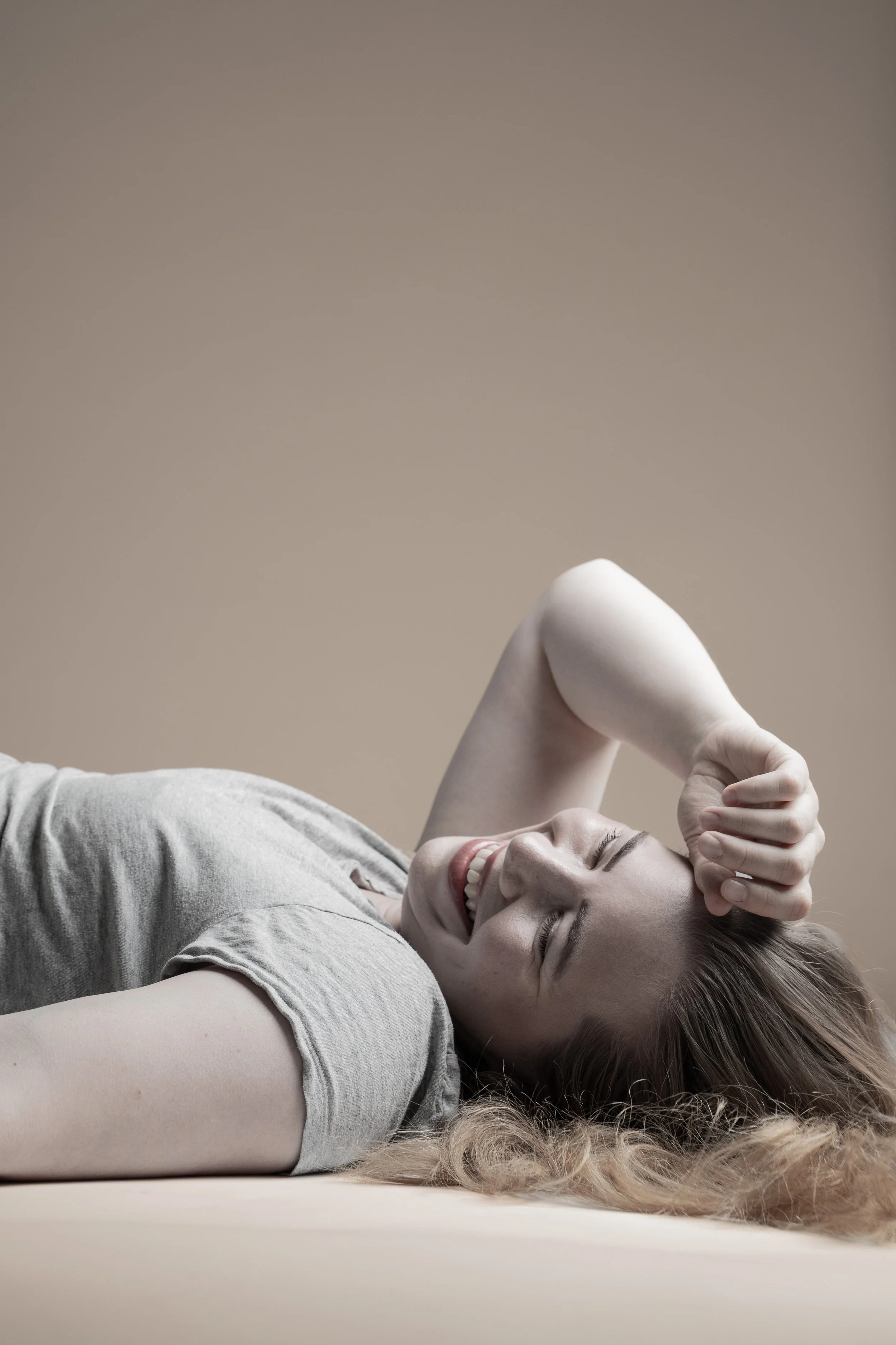 A woman with long, curly hair lying down on a beige surface, smiling with her eyes closed and hand resting on her forehead.