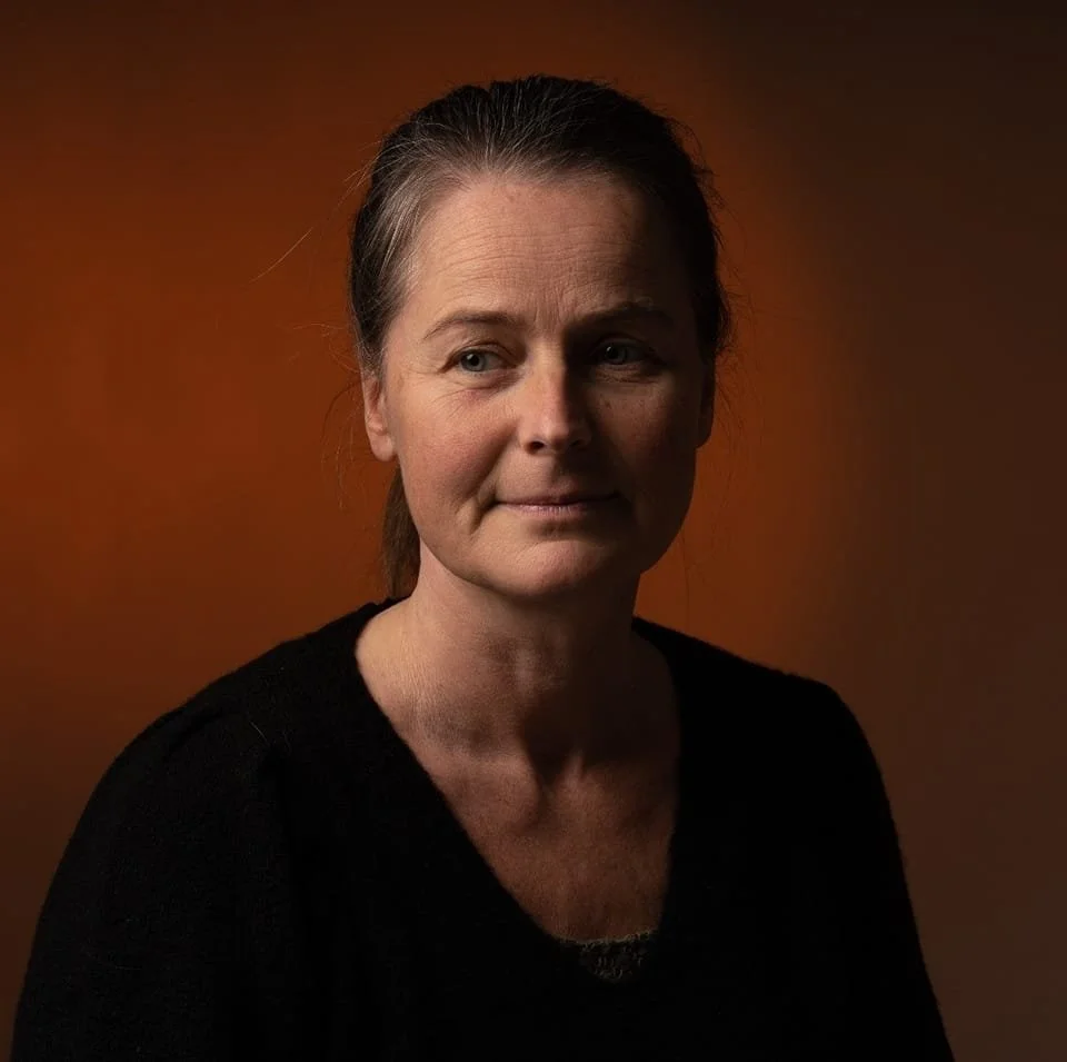 Portrait of a middle-aged woman with brown hair, wearing a black top, against a warm orange background.