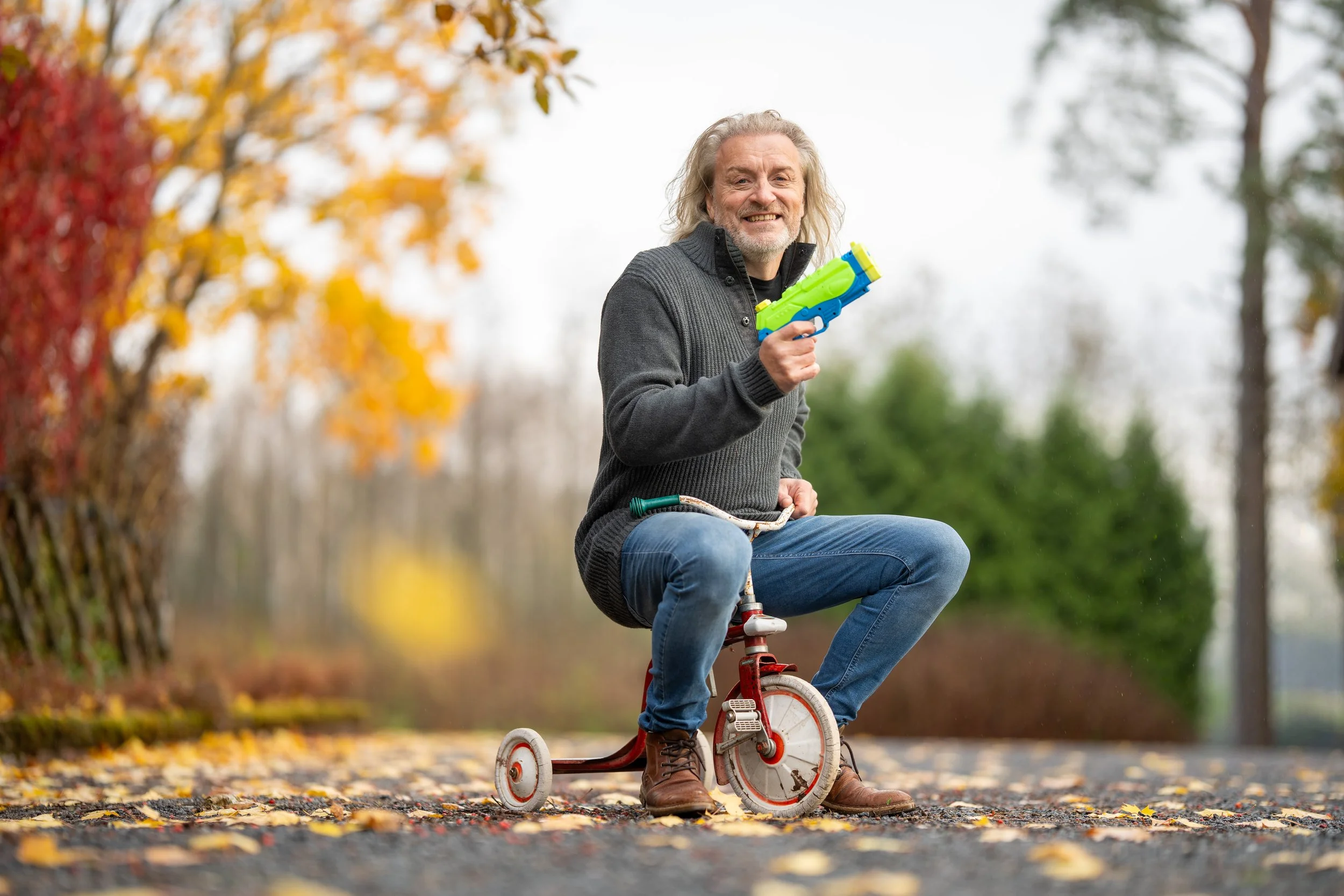 A cheerful man with curly gray hair and beard, wearing a dark sweater and jeans, sitting on a small red tricycle in an outdoor park with fall foliage, holding a colorful toy water gun.