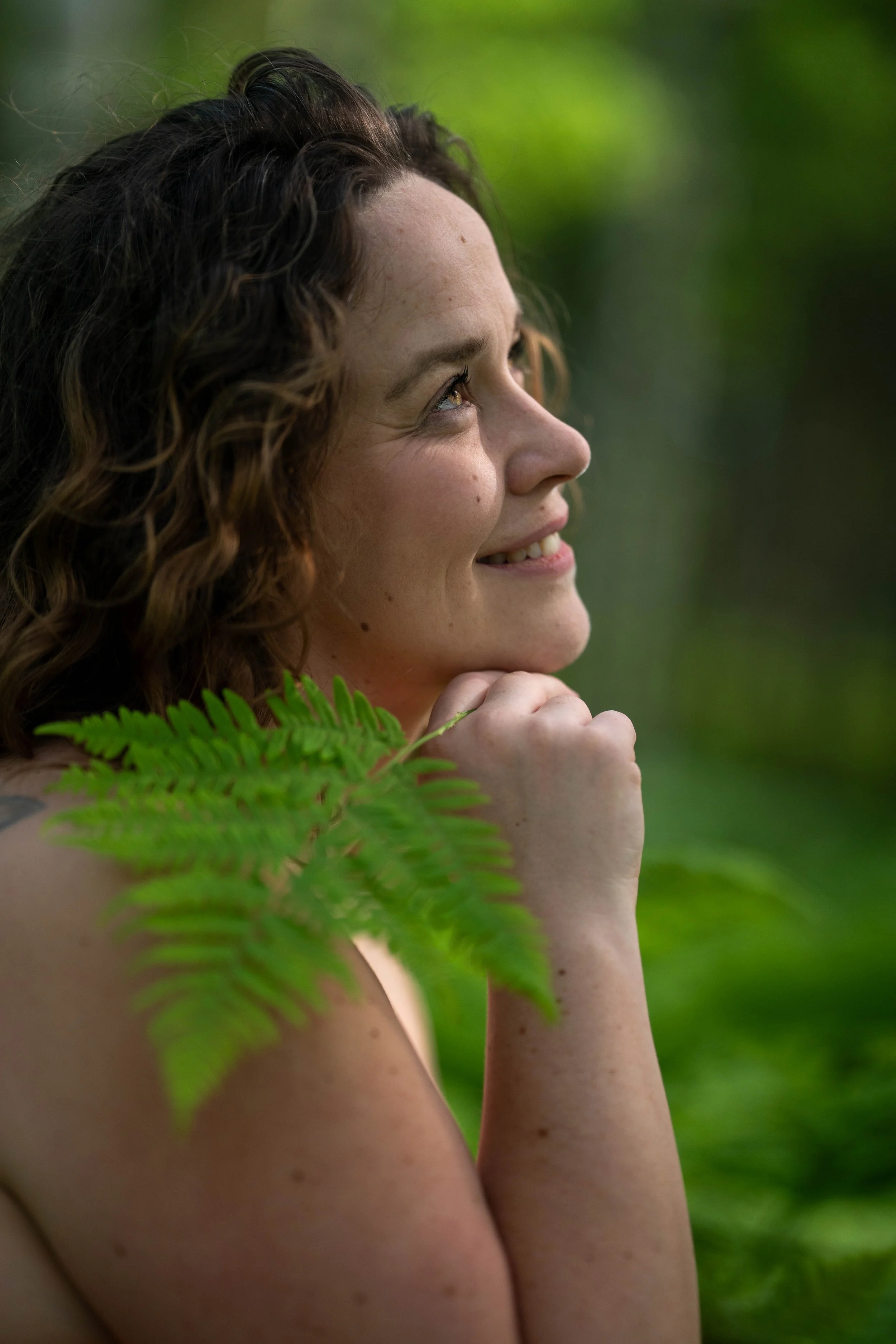 A woman with curly brown hair smiling in a green outdoor setting, resting her chin on her hand, with a fern leaf partially covering her shoulder.