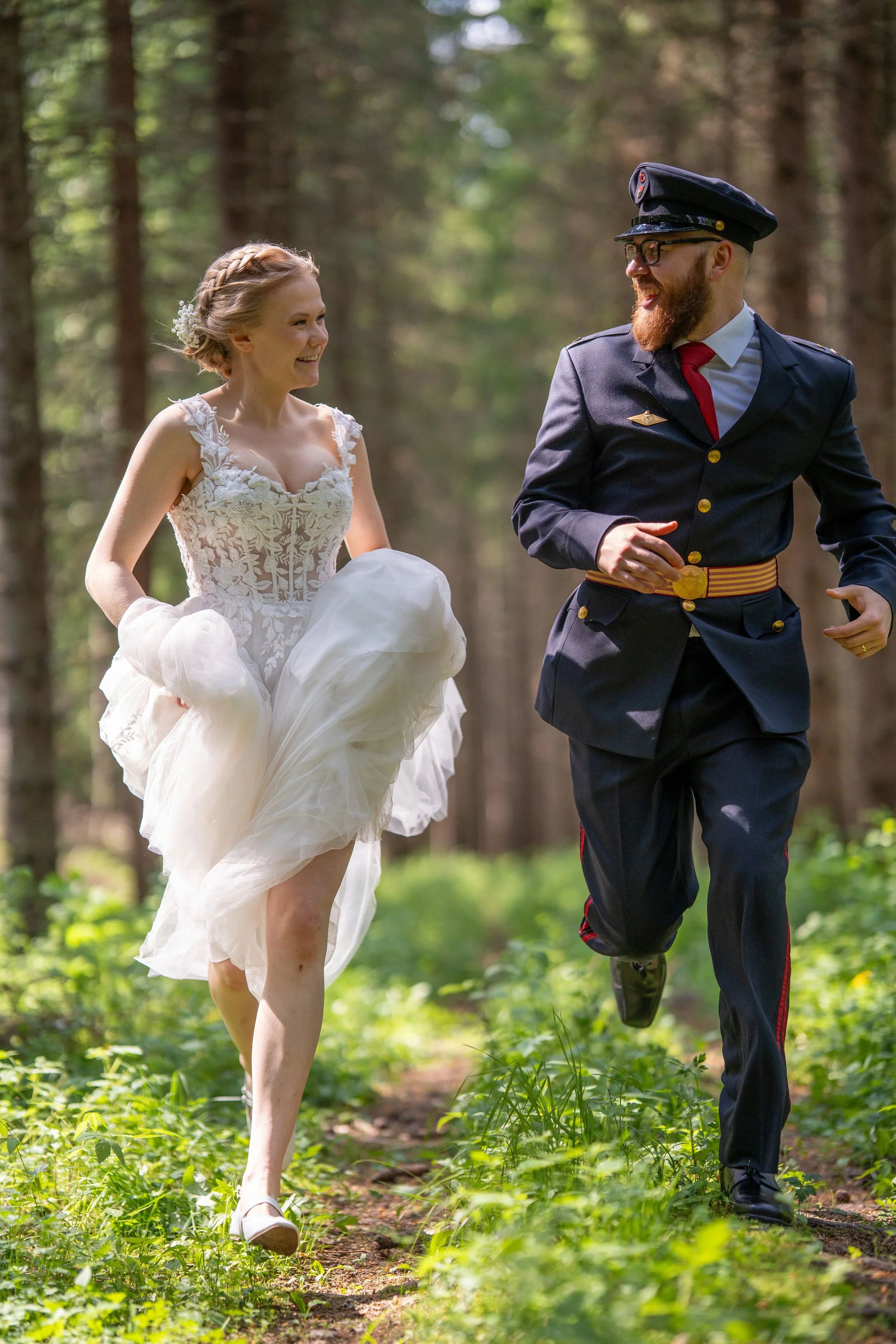 A bride in a white wedding dress and a groom in a military uniform running together through a forest.