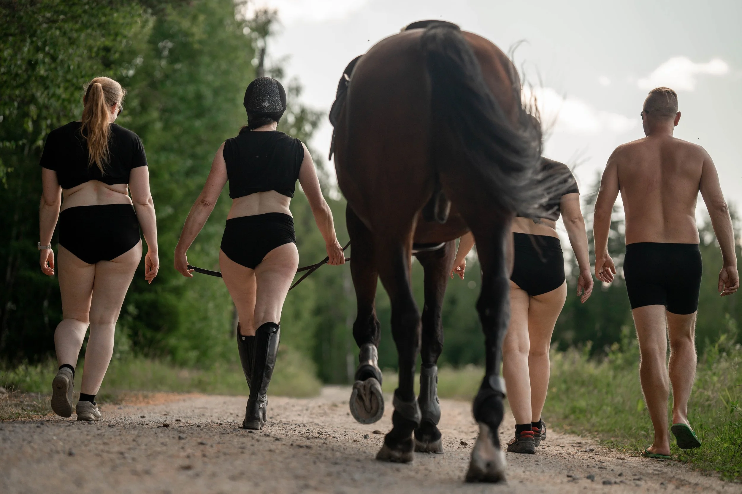 Five people, dressed in minimal clothing, walking on a dirt path in a forested area, led by a person riding a horse. The scene appears to be outdoors during daytime.