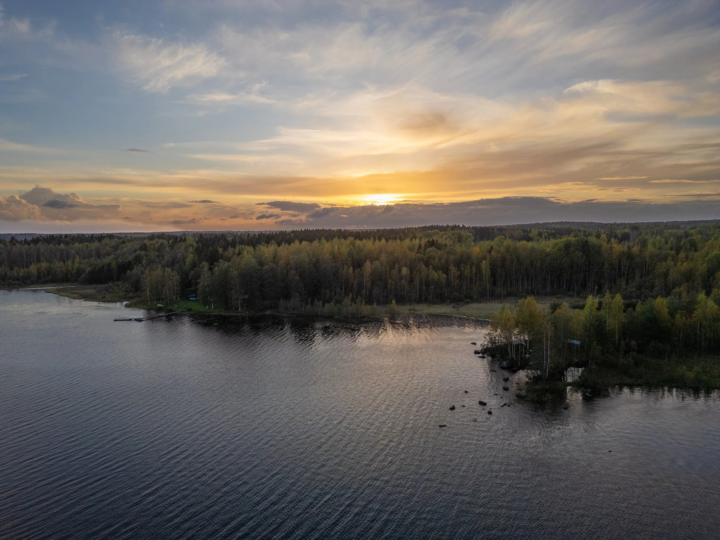 Aerial view of a river with a tree-lined shore at sunset, with a partly cloudy sky and the sun near the horizon.