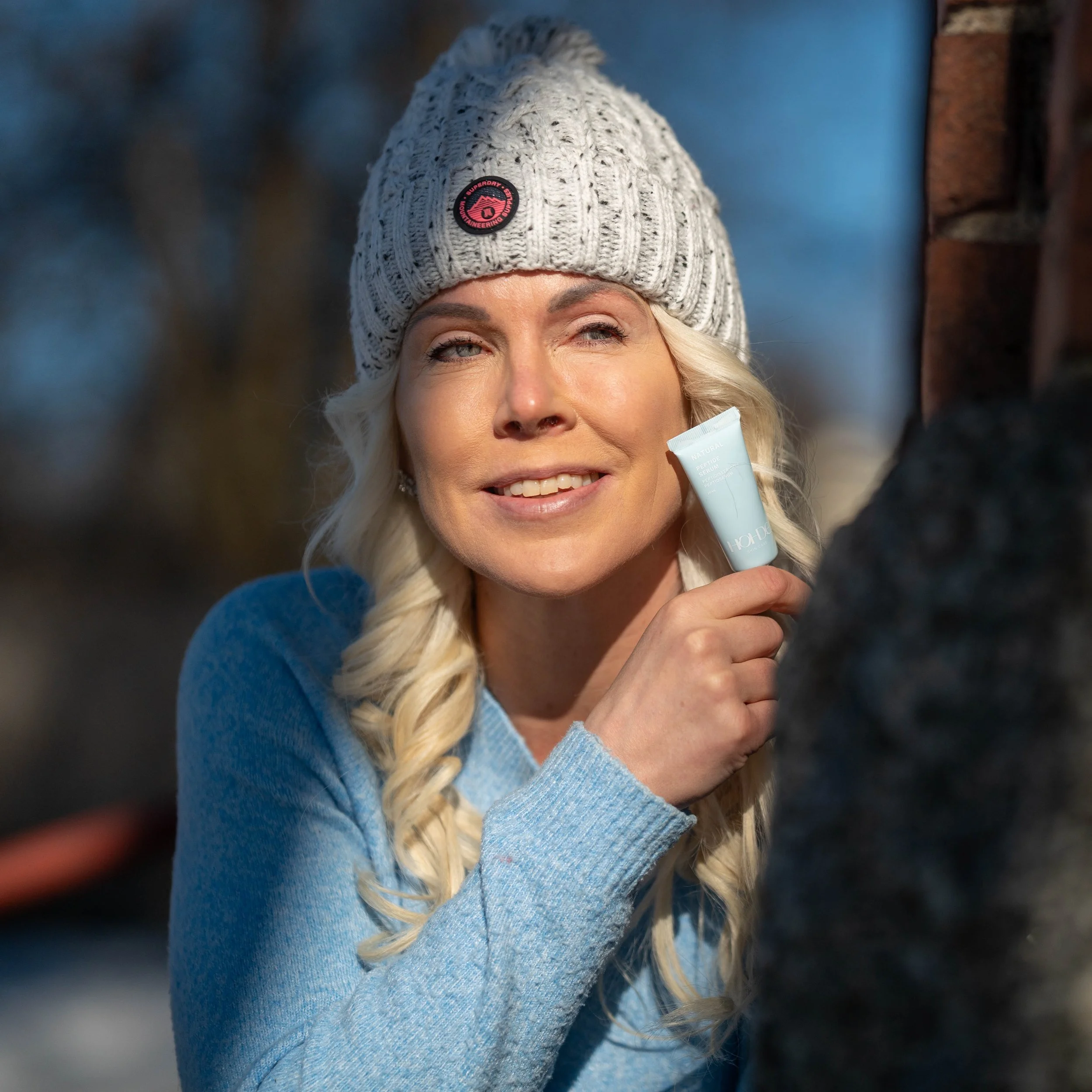 A woman with blonde hair in a braid, wearing a gray knit beanie and a light blue sweater, holds a small tube of cream near her face outdoors. She is smiling at the camera with a brick wall and blue sky in the background.