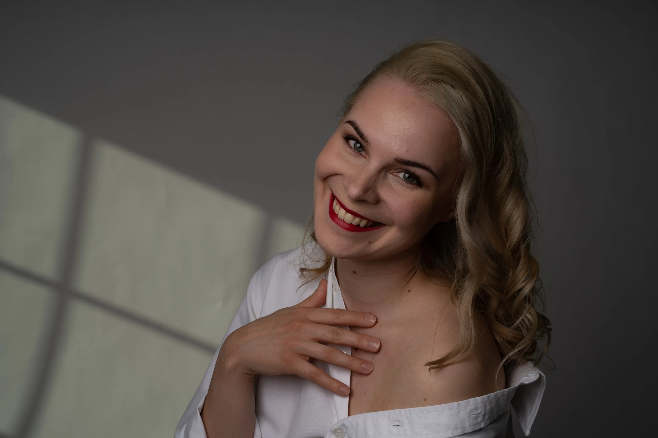 Smiling woman with blonde hair and red lipstick, wearing a white shirt, touching her chest with her right hand, against a gray background with a shadow.