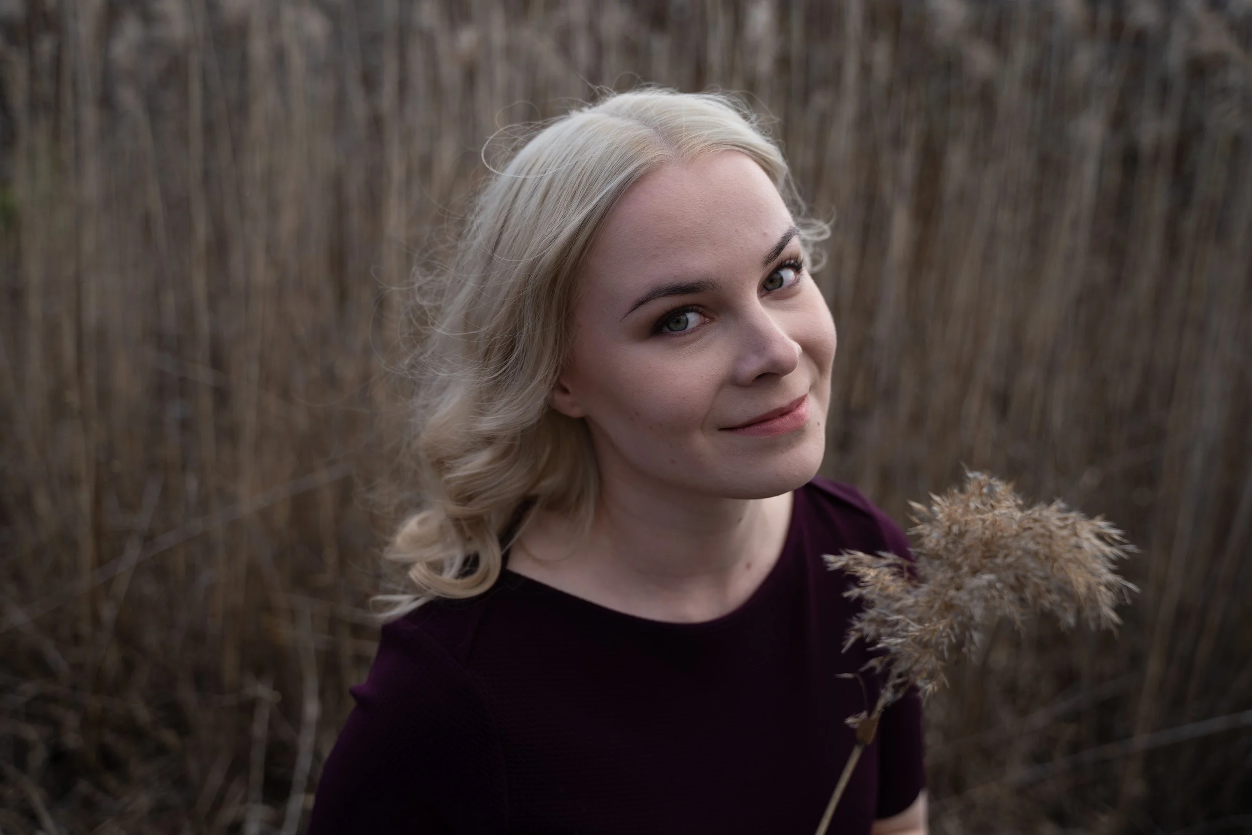 A woman with curly blond hair and a maroon top holding a dried plant in a field of tall, brown grass, smiling at the camera.