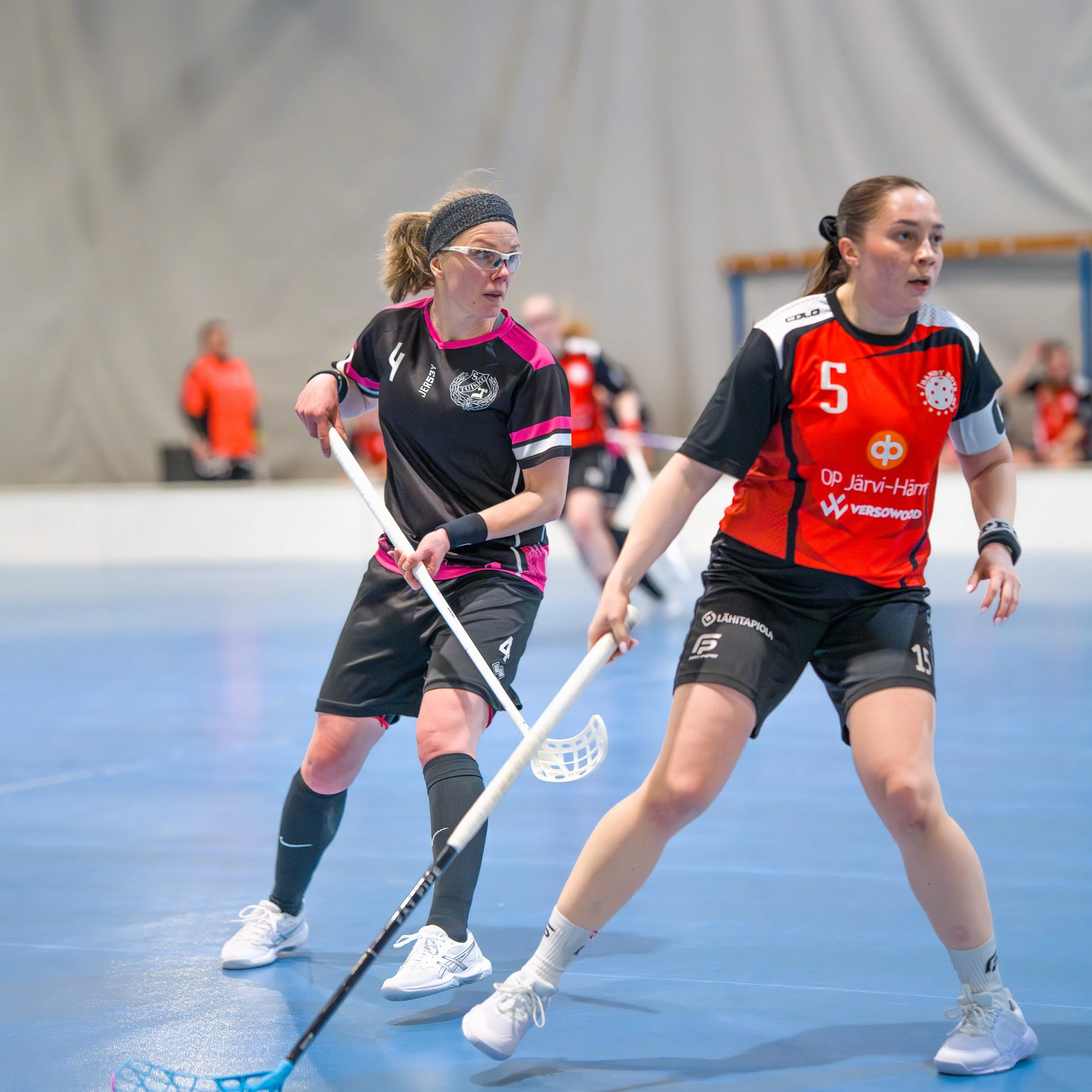 Two women playing indoor floor hockey, one in a black and pink jersey and the other in a red and black jersey, on a blue court.
