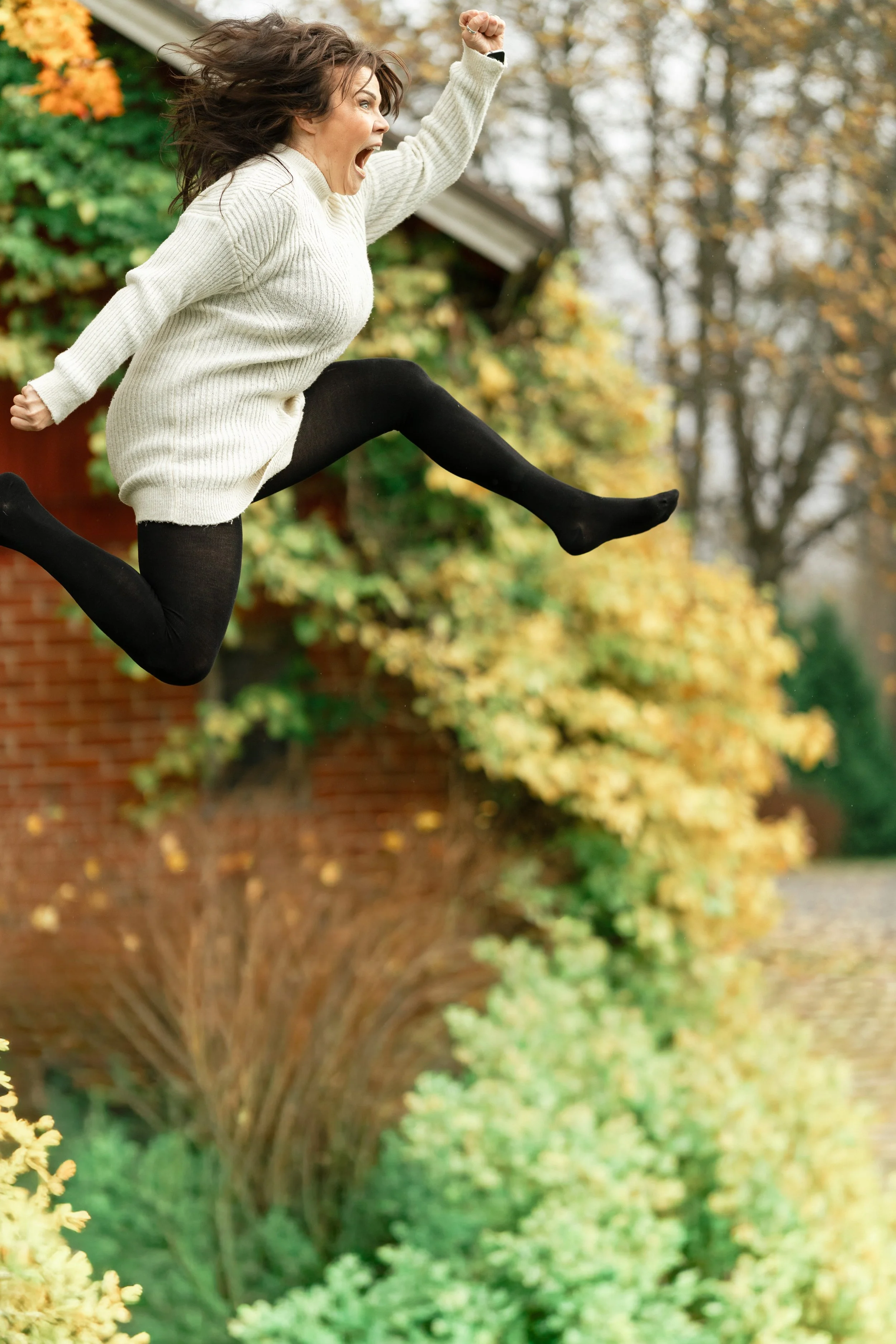 A woman joyfully jumping outdoors during autumn with trees and a house in the background.