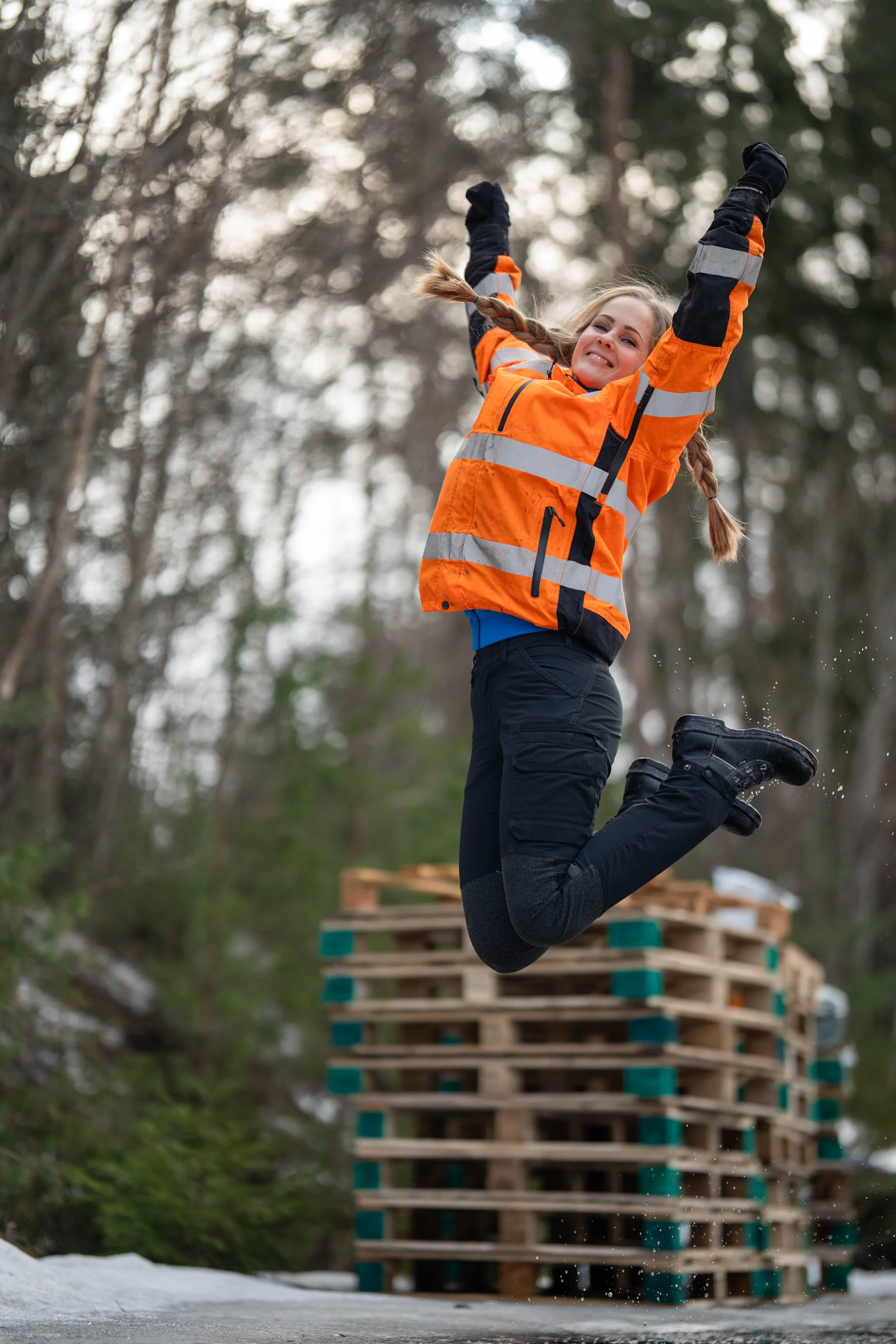 A woman wearing an orange safety jacket jumping in the air outdoors near wooden pallets with a forest background.