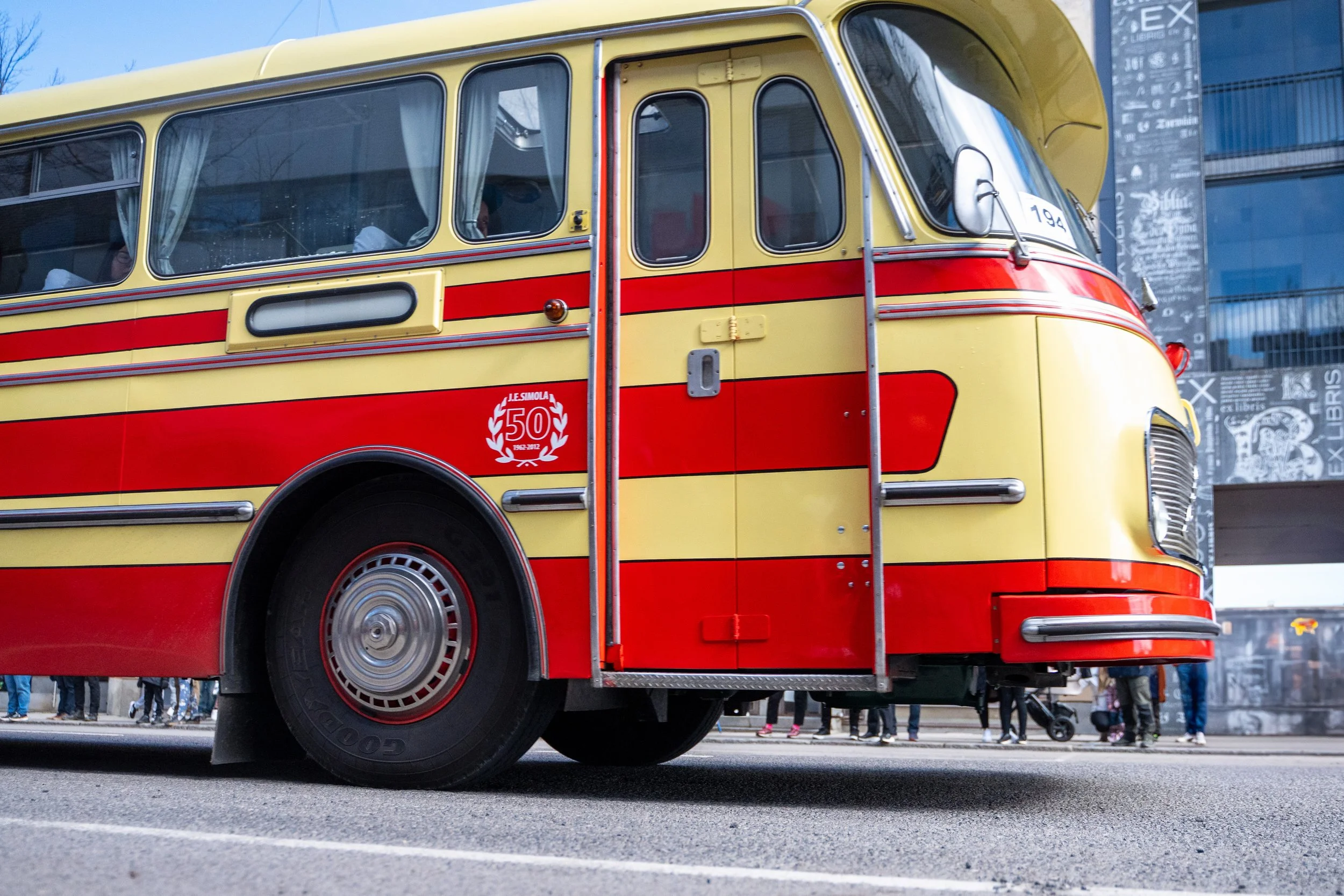 A vintage yellow and red bus, marked with a 50th anniversary badge, driving on a city street with pedestrians in the background.