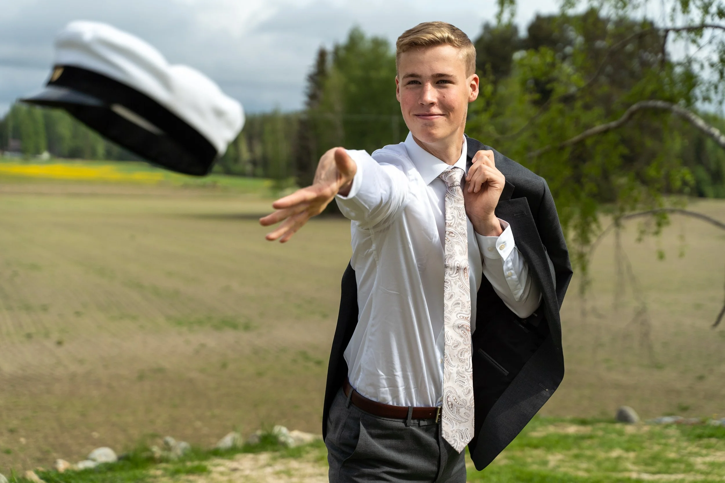 A young man in a formal white shirt, tie, and gray suit jacket carrying another gray suit jacket over his shoulder, throws a graduation cap into the air outdoors on a cloudy day.