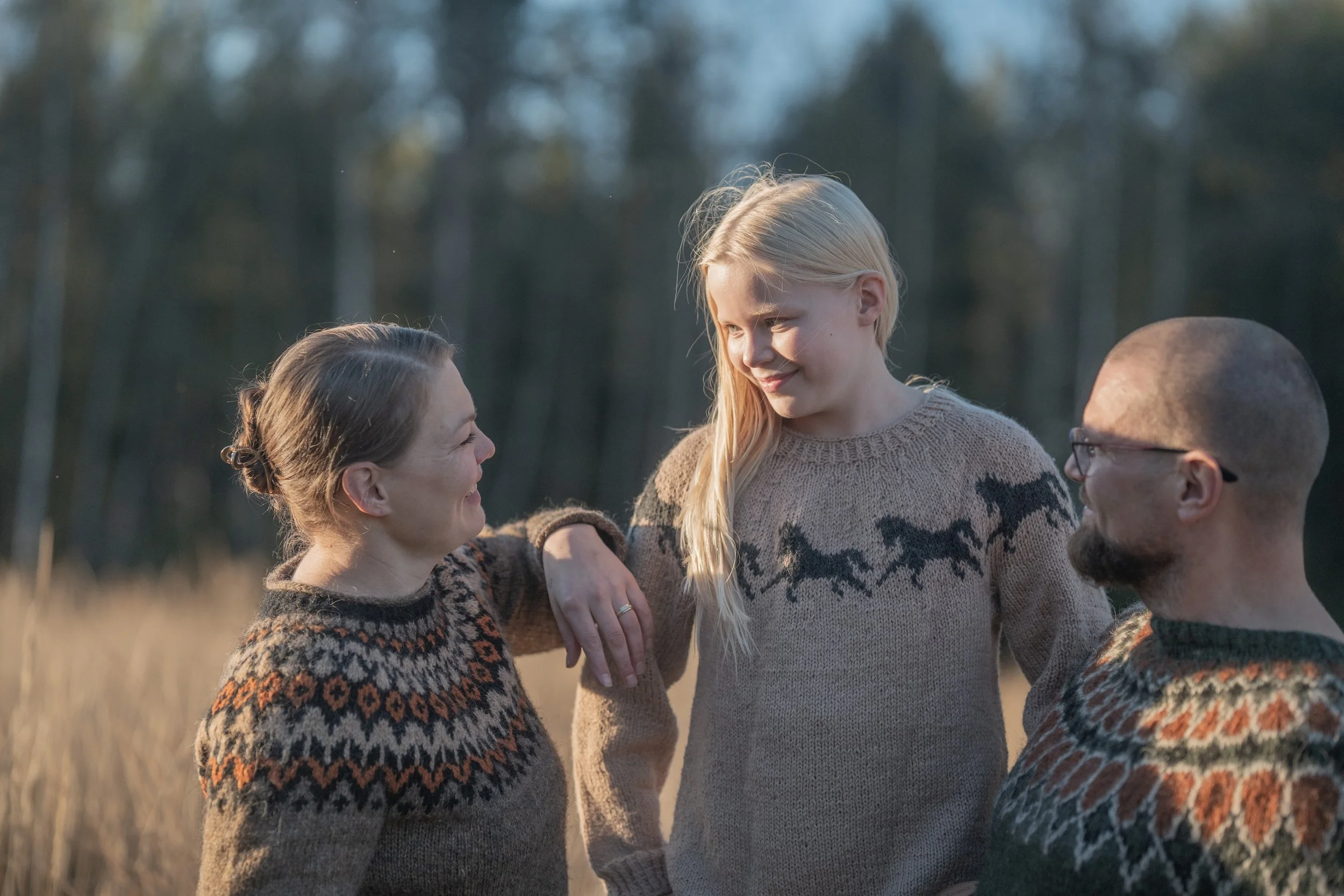 A family of three outdoors in a field during daylight, all wearing sweaters with Nordic patterns, engaging in a warm, happy conversation.