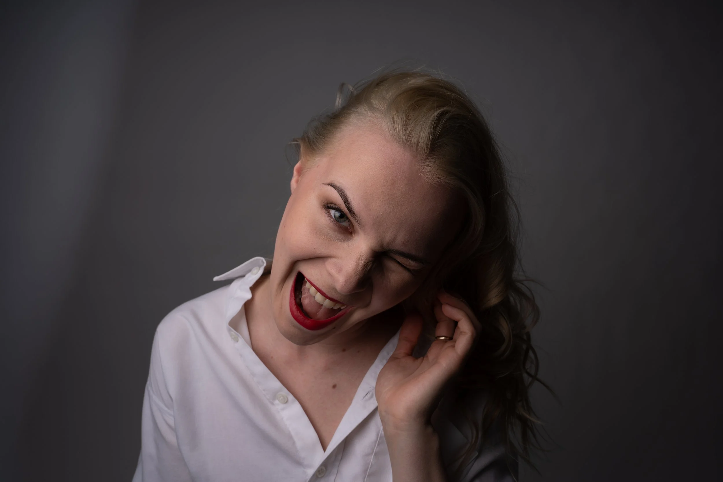 A woman with blonde hair, wearing a white shirt, smiling and winking at the camera, with her hand near her ear against a dark gray background.