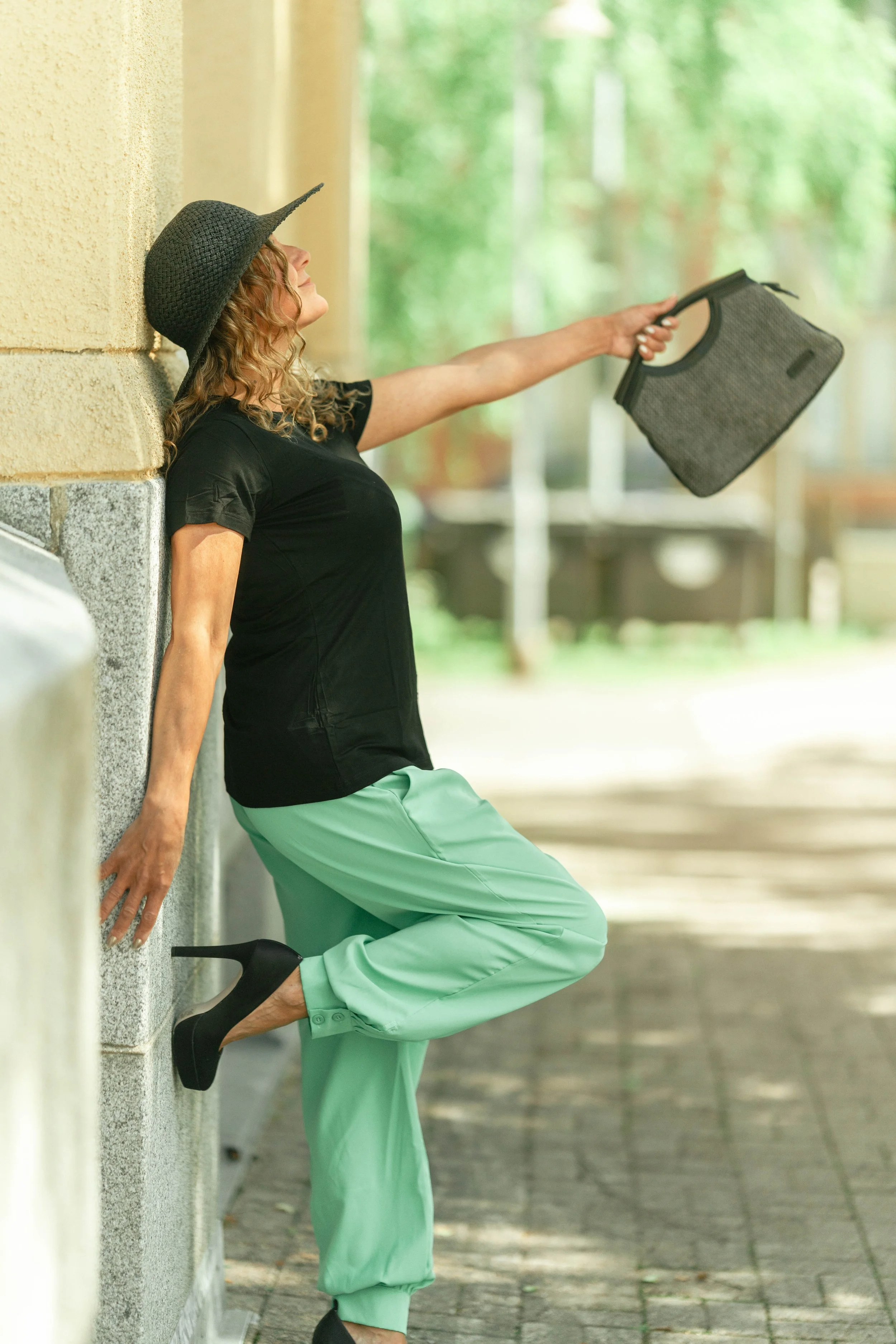 A woman with curly hair, wearing a black hat, black shirt, mint green pants and black high heels, leaning against a wall and reaching out with her arm extended holding a grey handbag.