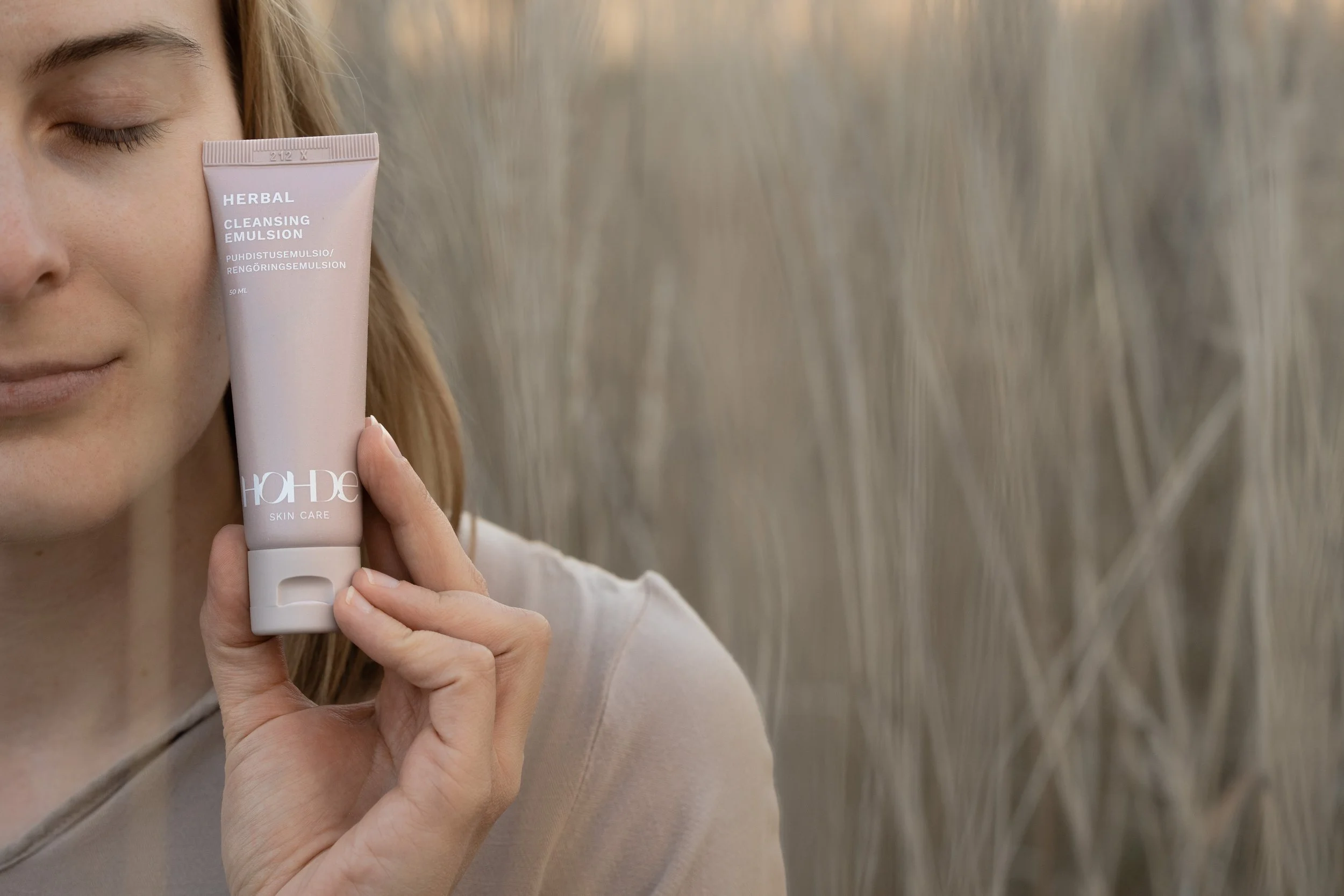 A woman in a beige top holding a pink tube of Herbal Cleansing Emulsion close to her face outdoors, with tall dry grass in the background.