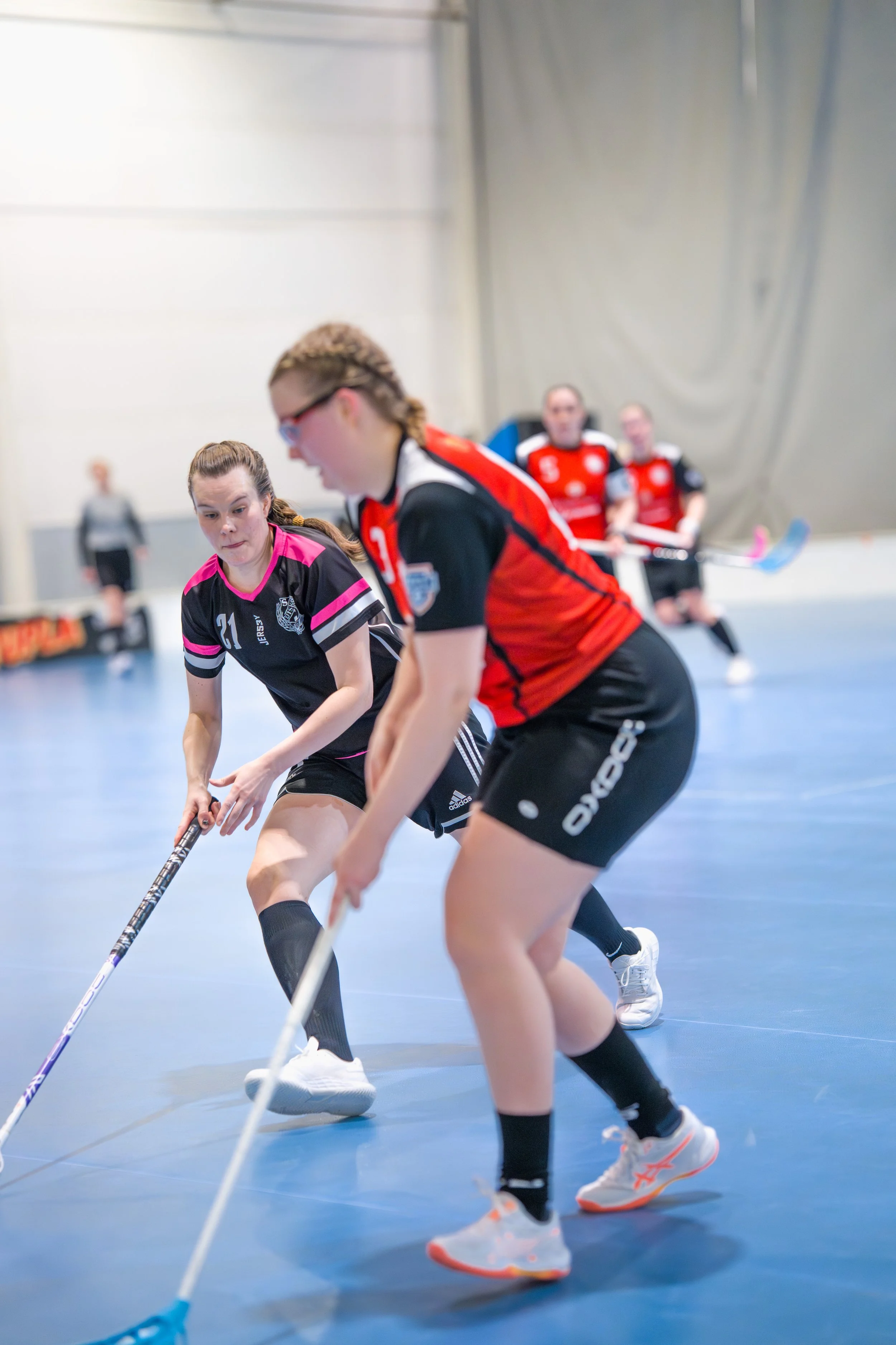 Women playing indoor floor hockey on a blue court, one woman in a black and pink jersey and another in a black and red jersey, with others in red jerseys in the background.