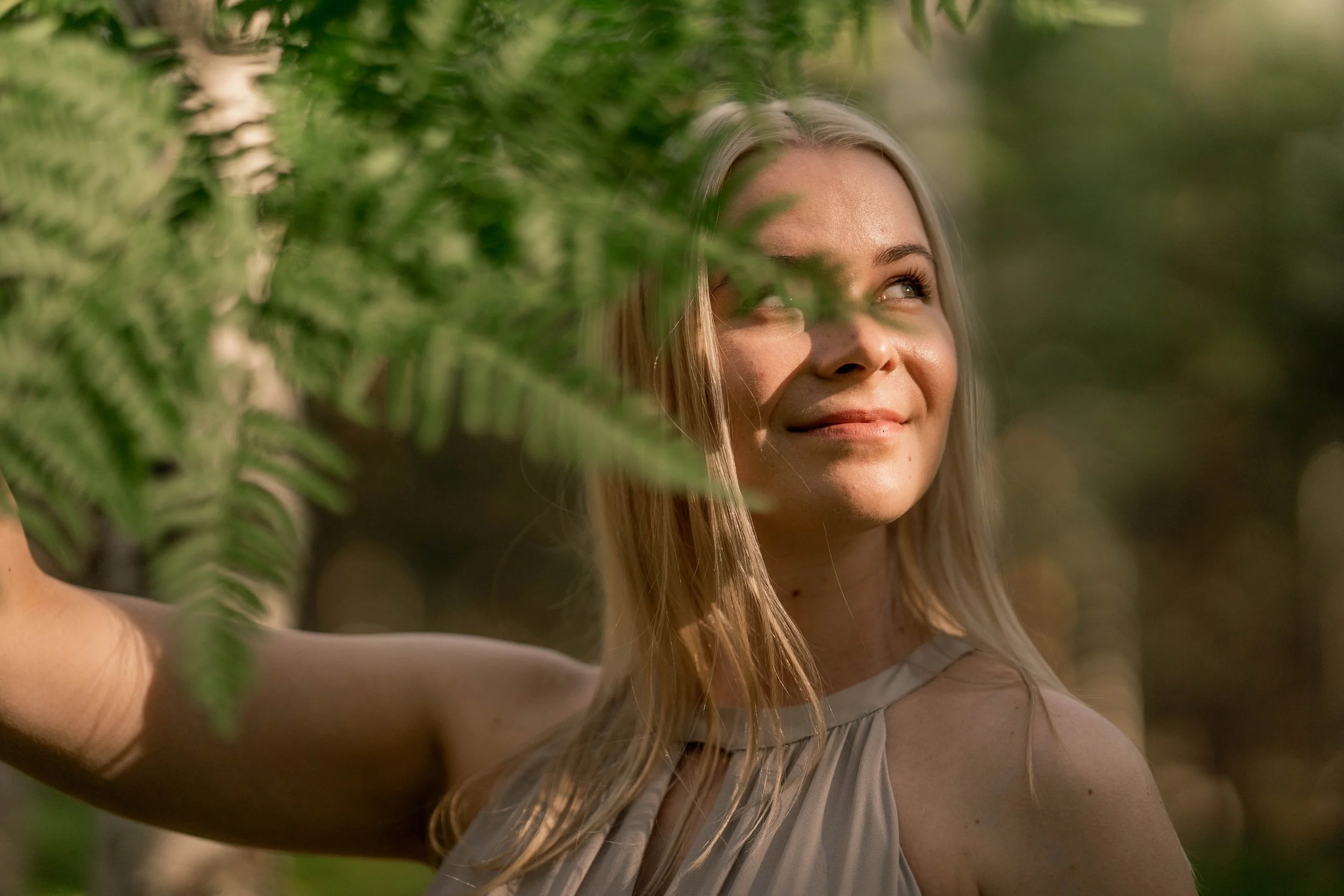 A woman with blonde hair smiling in a forest, partially obscured by green fern leaves.