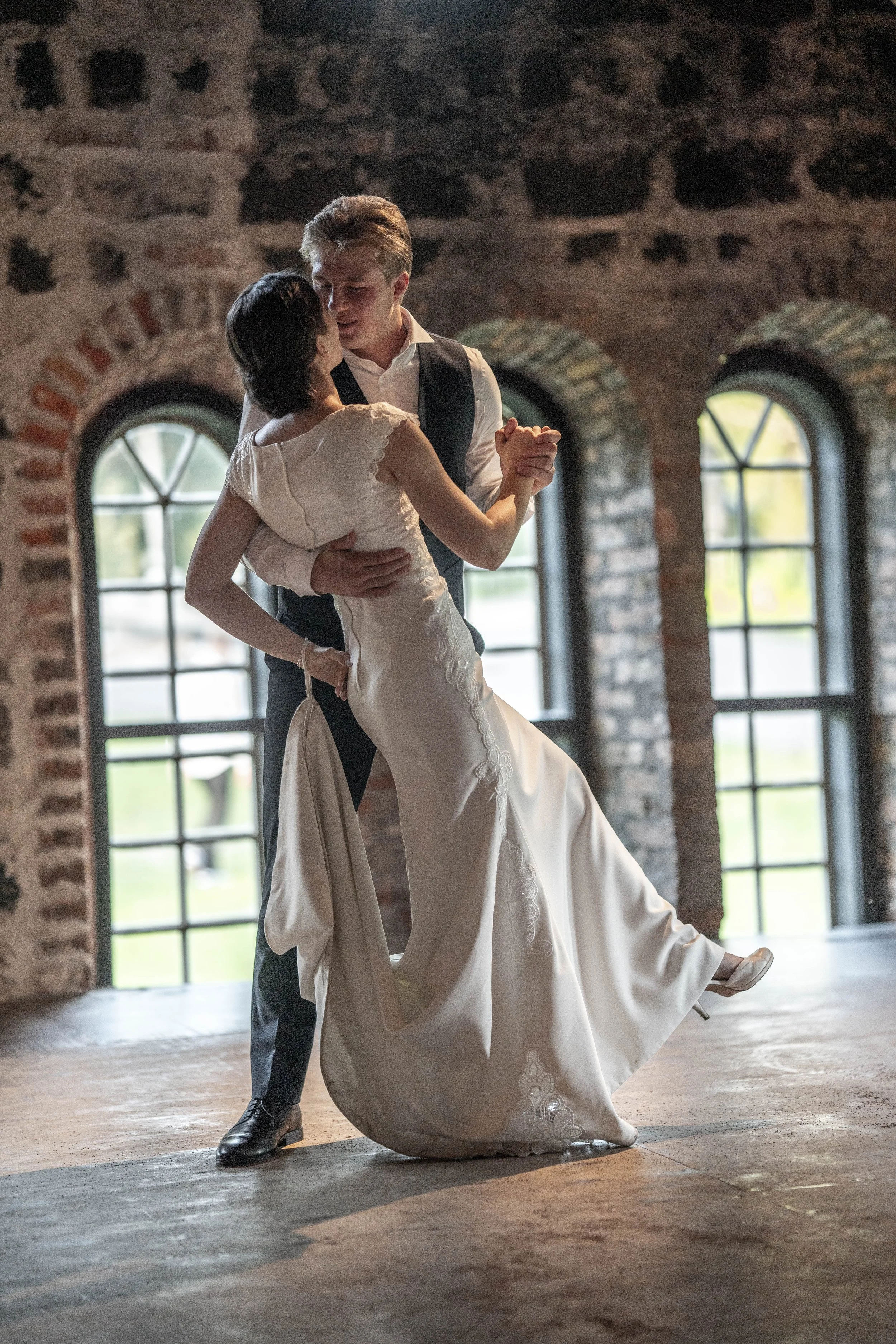 A young couple dancing closely in a rustic indoor setting with large arched windows.