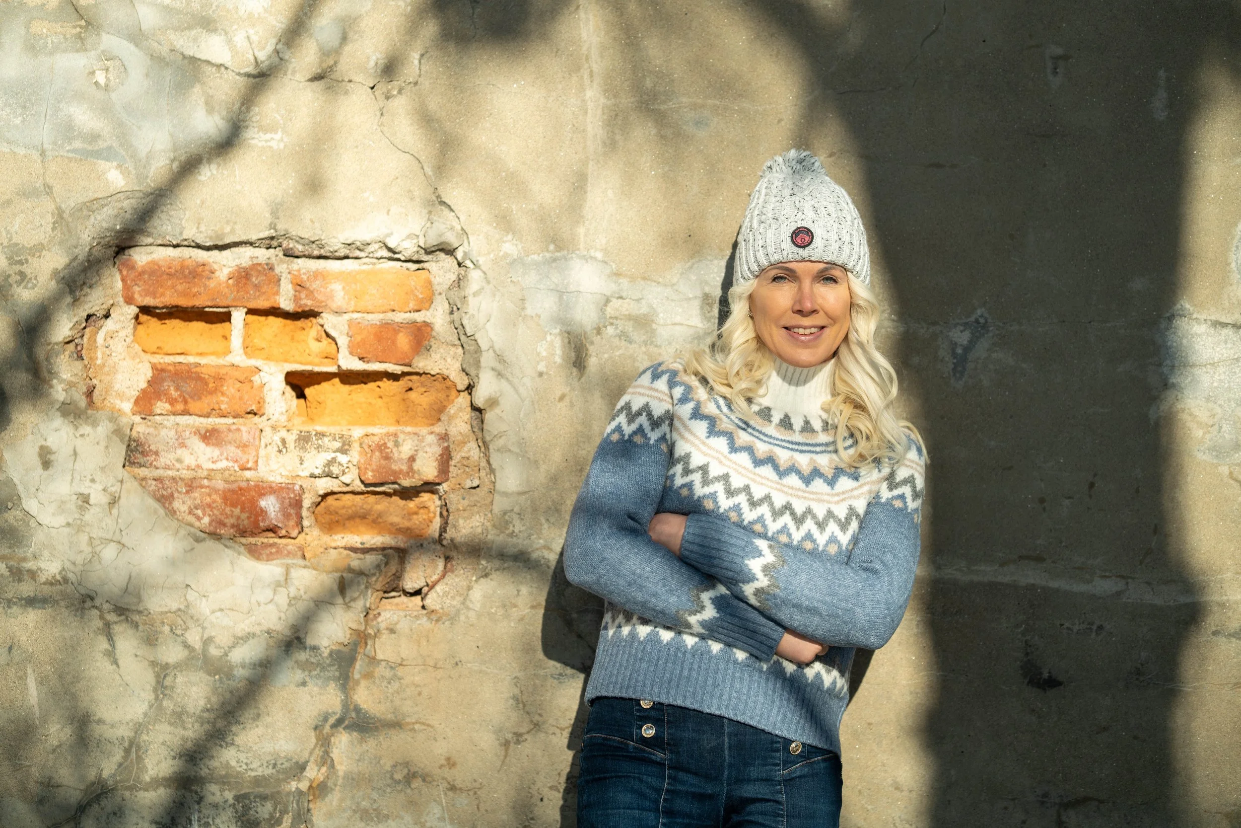 A smiling woman with blonde hair, wearing a gray knit hat and a blue and white patterned sweater, standing with arms crossed against a weathered concrete wall with exposed red bricks.