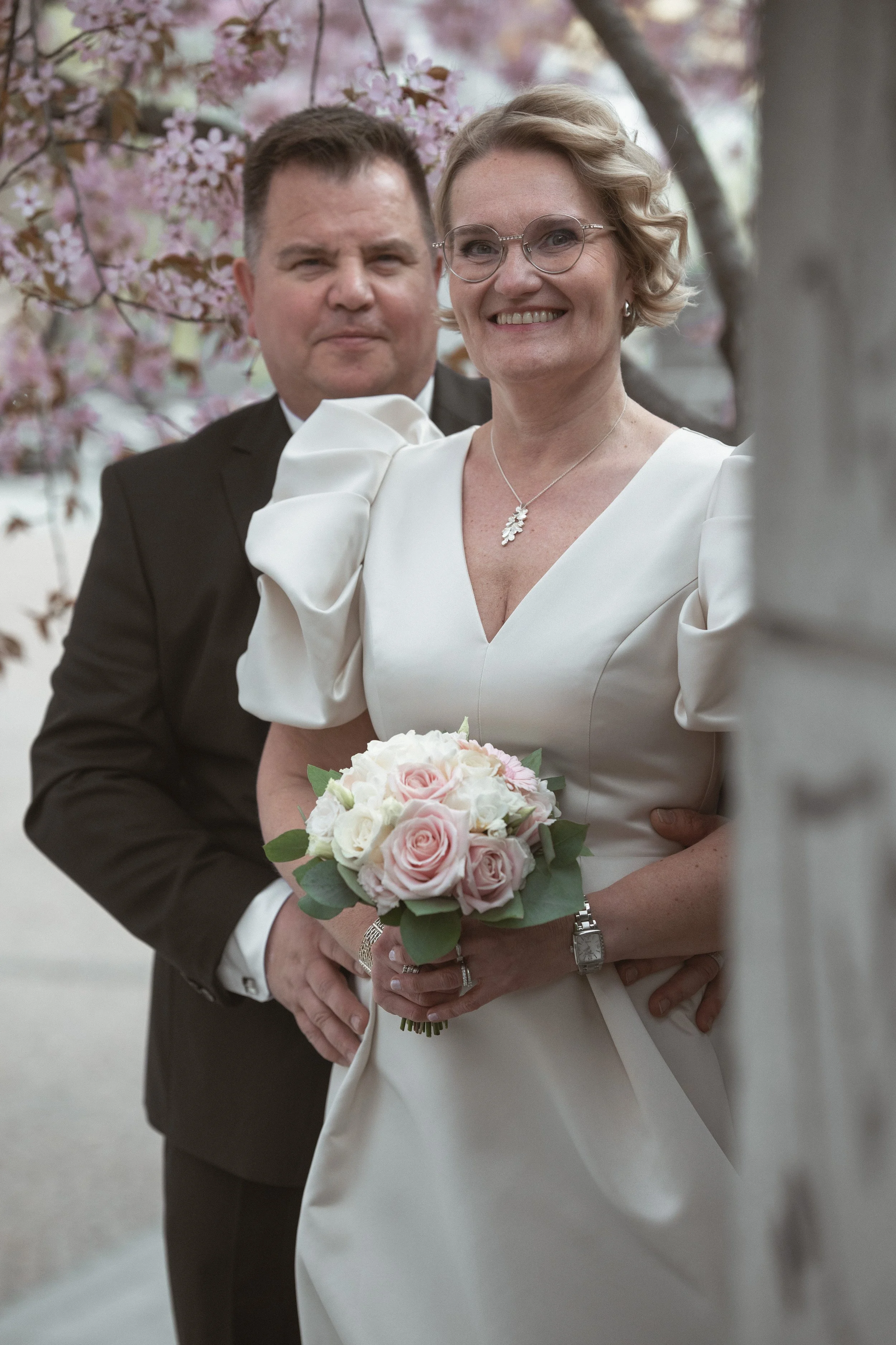 A smiling woman in a white wedding dress holding a bouquet of pink and white roses stands in front of a man in a black suit, with pink cherry blossom trees in the background.