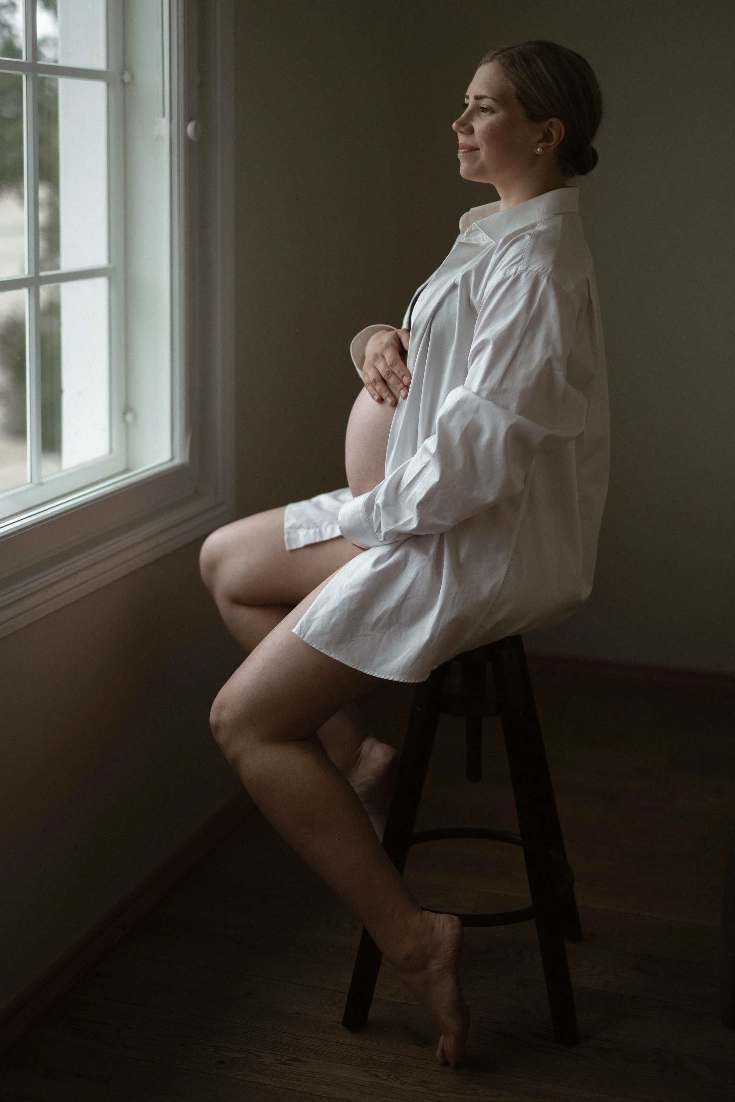 A pregnant woman wearing a white shirt sitting on a stool by a window, gazing outside with a peaceful expression.