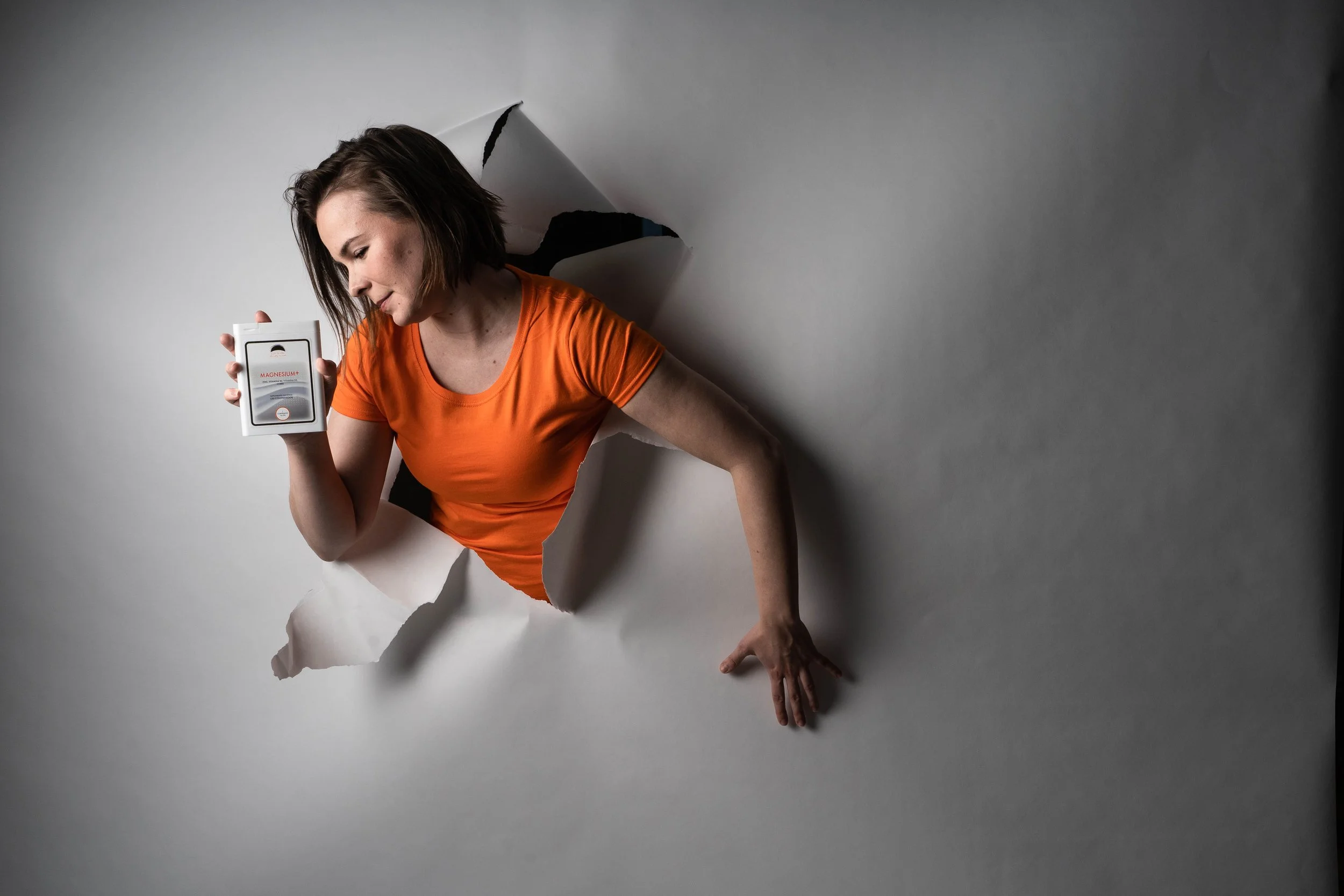 A woman in an orange shirt is emerging through a large hole in a white paper backdrop, holding a box of magnesium supplement in her right hand.