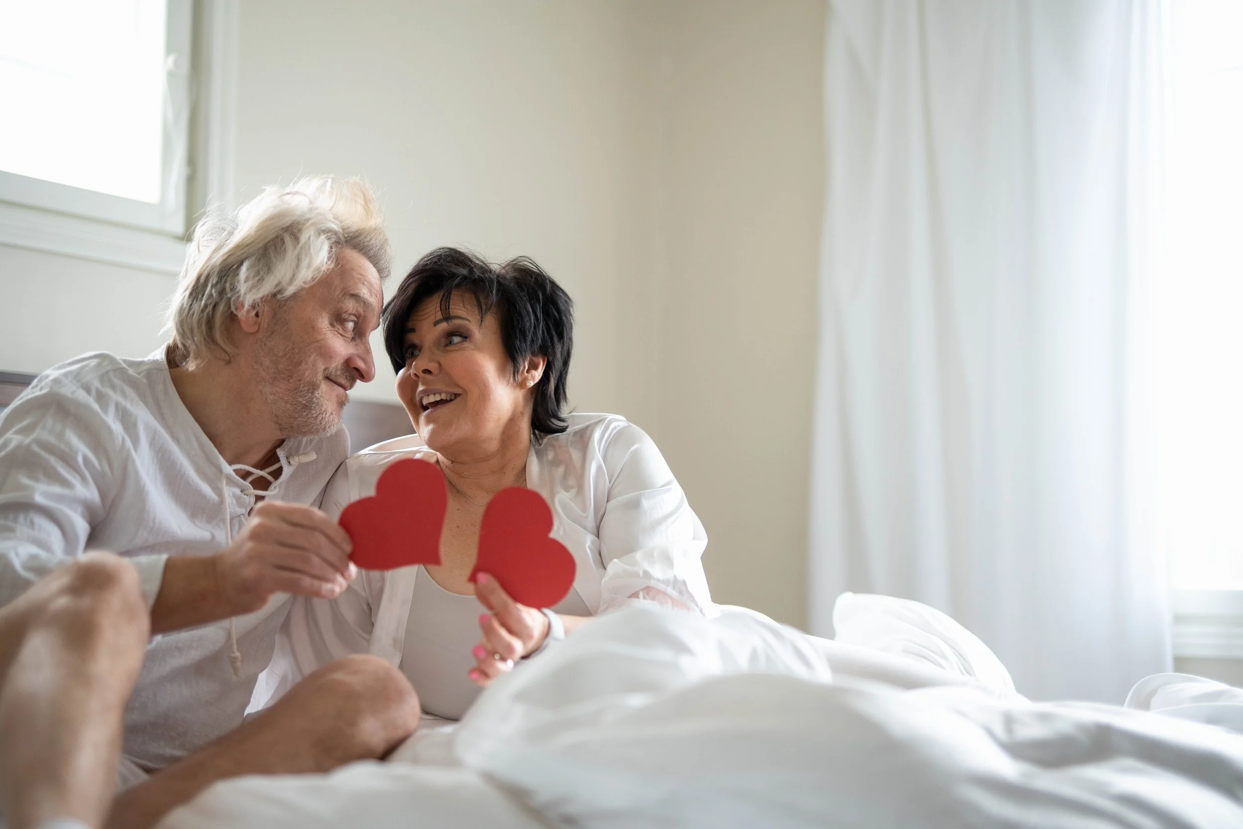 An elderly couple sitting on a bed, smiling at each other, holding red paper hearts in a bright bedroom with white curtains.