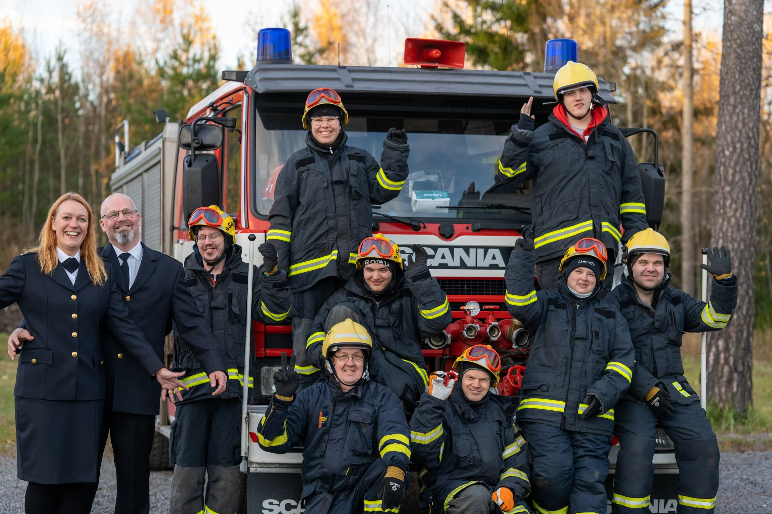 Group of firefighters, some in uniform with helmets and others in formal uniforms, posing in front of a fire truck outdoors during daytime with autumn trees in the background, smiling and waving.