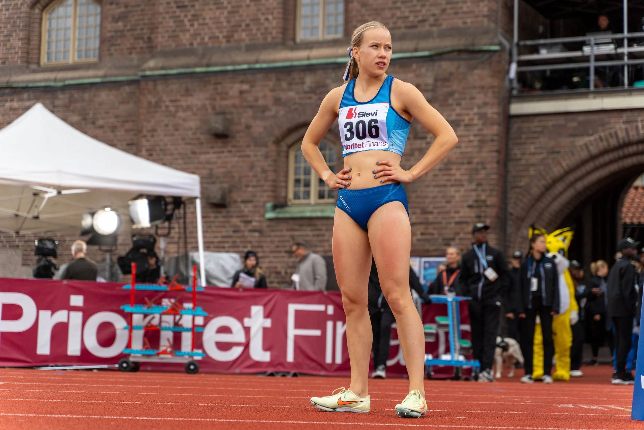 A female athlete standing on a track with her hands on her hips, wearing a blue sports bra and shorts, with a competition bib number 306, at an outdoor track event.