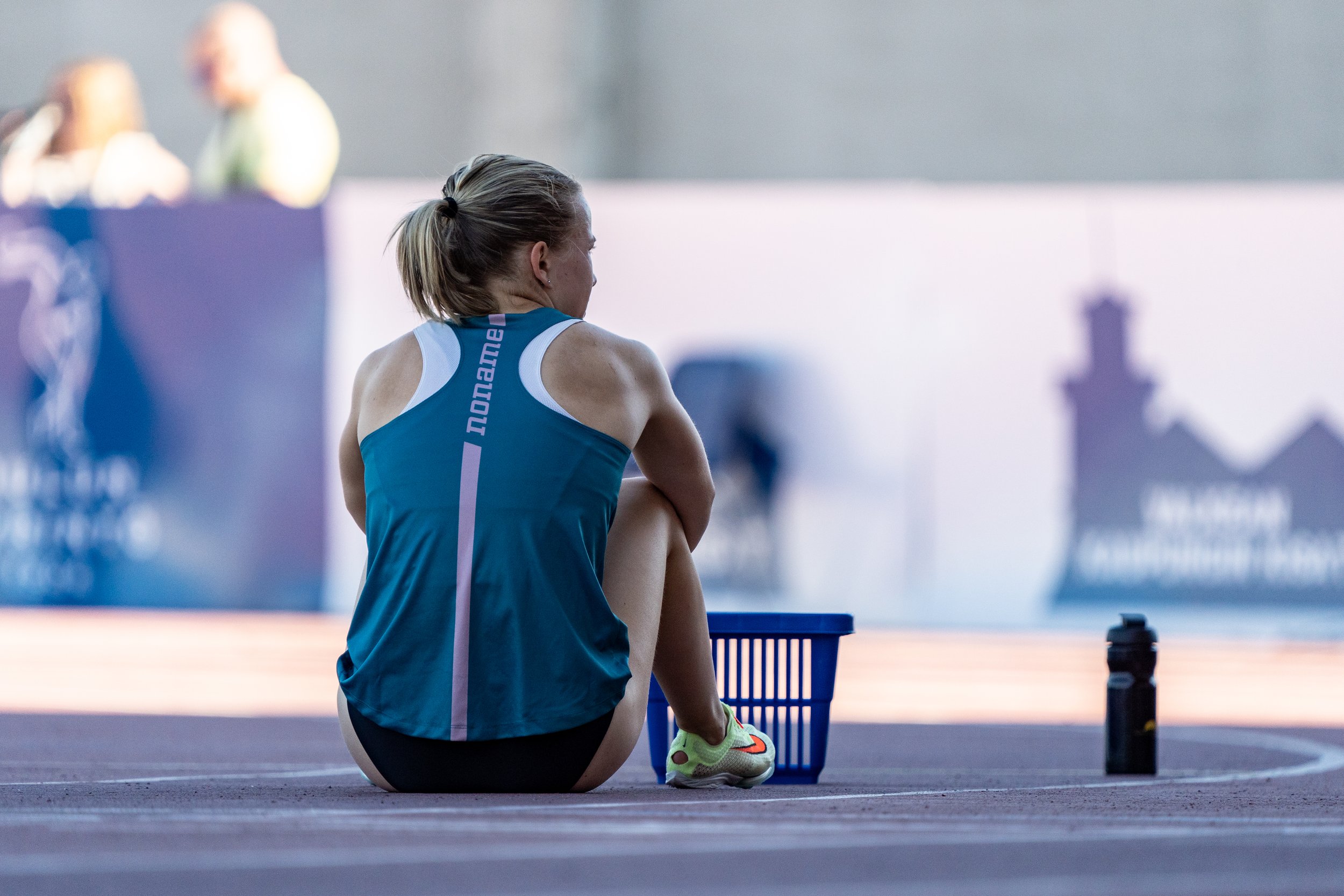 A female athlete sitting on a track next to a blue basket and a black water bottle, wearing a blue sports jersey, with her hair in a ponytail.