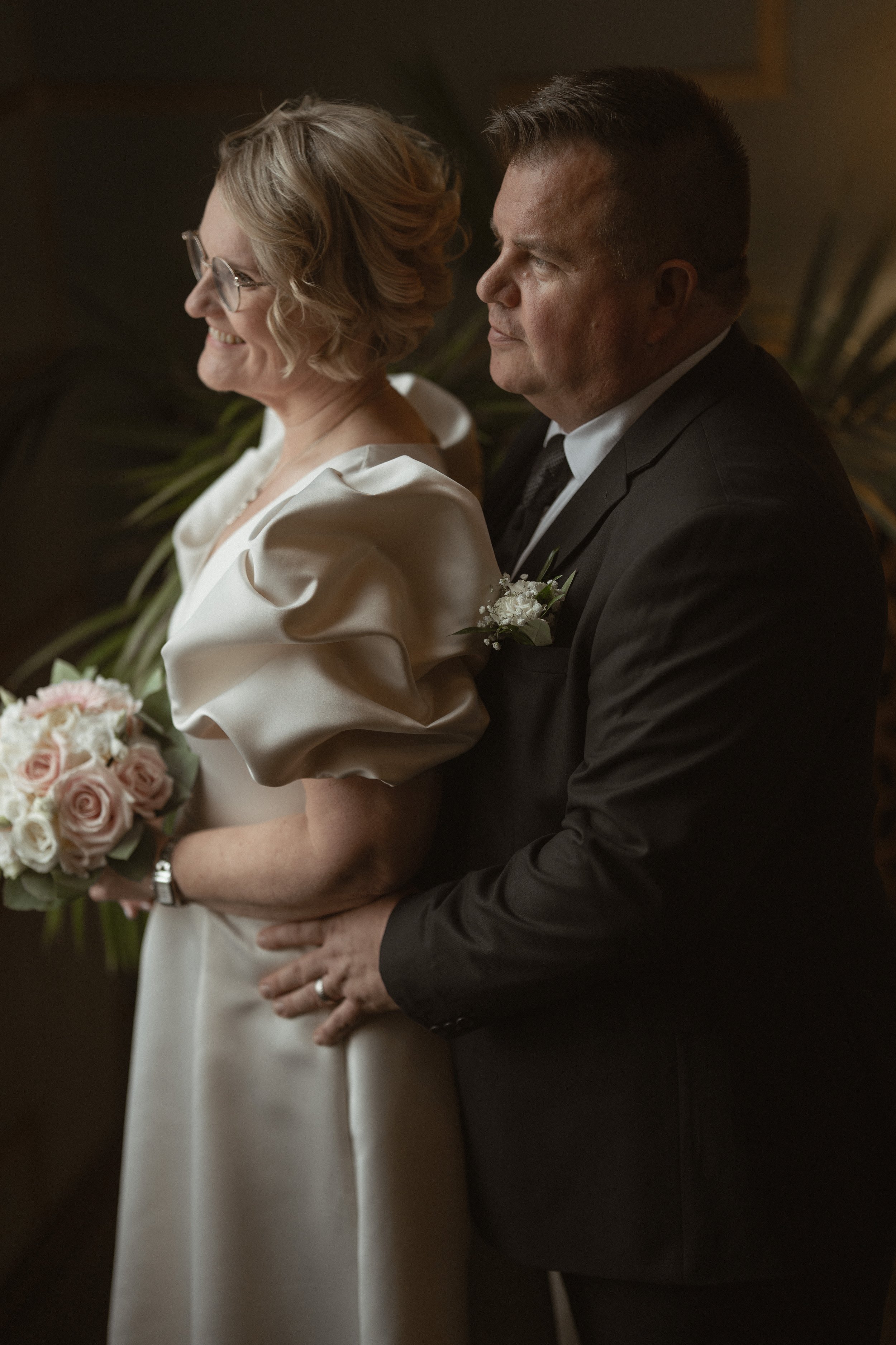 A bride and groom hold each other during their wedding ceremony. The bride is smiling and holding a bouquet of pink and white roses, wearing a satin dress with puffed sleeves. The groom is in a black suit and boutonniere, standing behind her with his