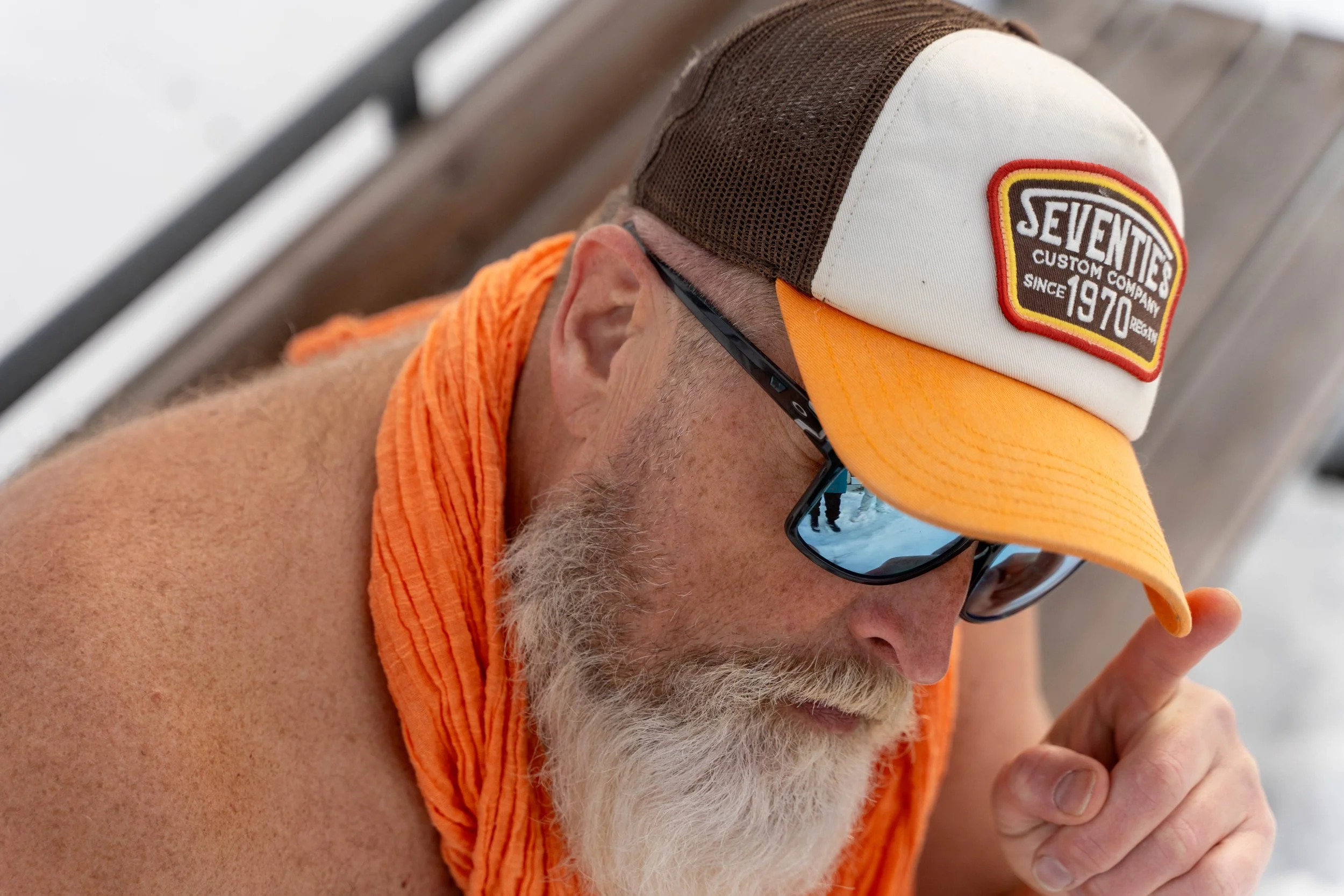 A man with a white beard wearing a multicolored cap, sunglasses, and an orange sleeveless shirt or towel, sitting on a bench.