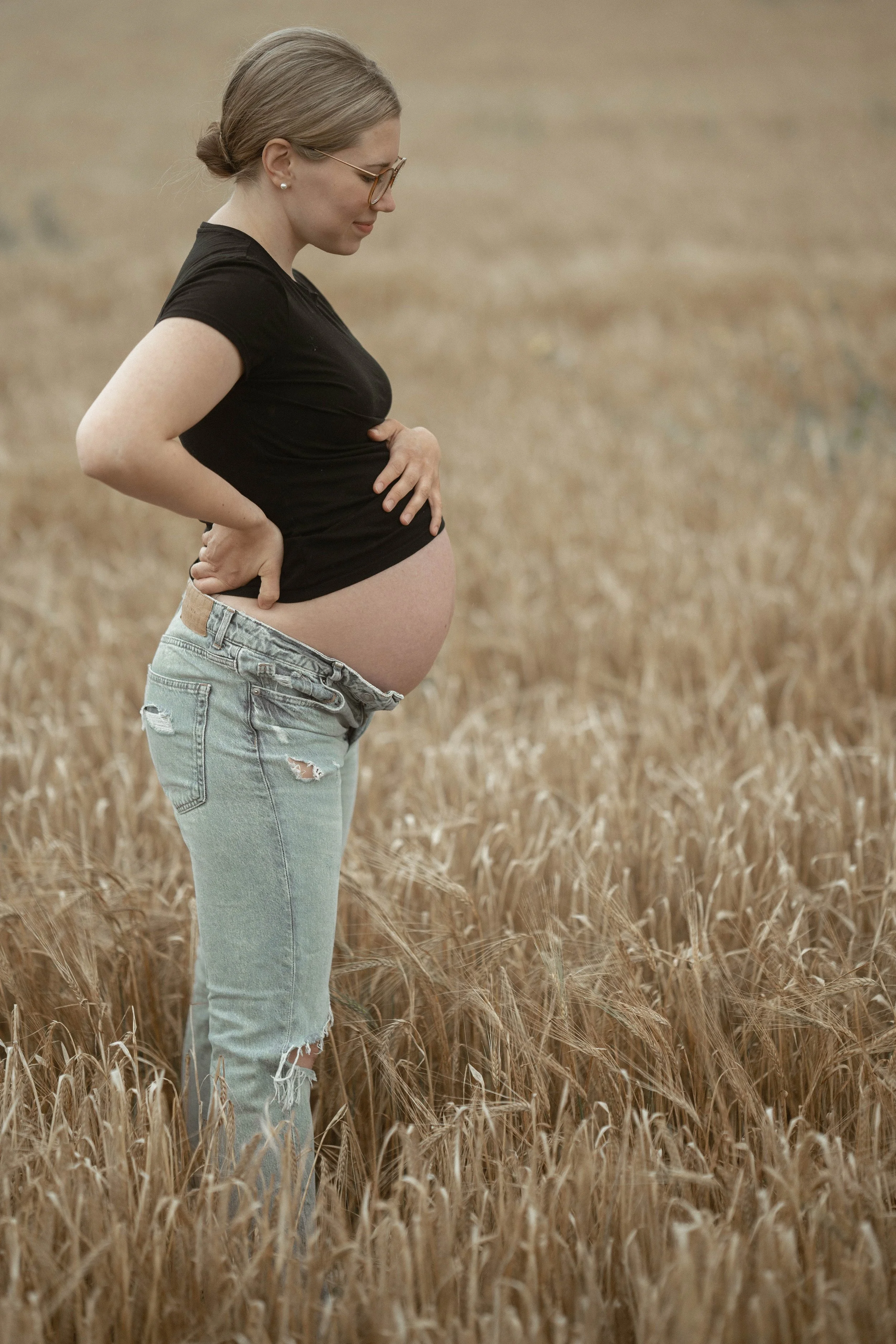 Pregnant woman standing in a wheat field, touching her belly with her hands, wearing a black t-shirt and ripped jeans.