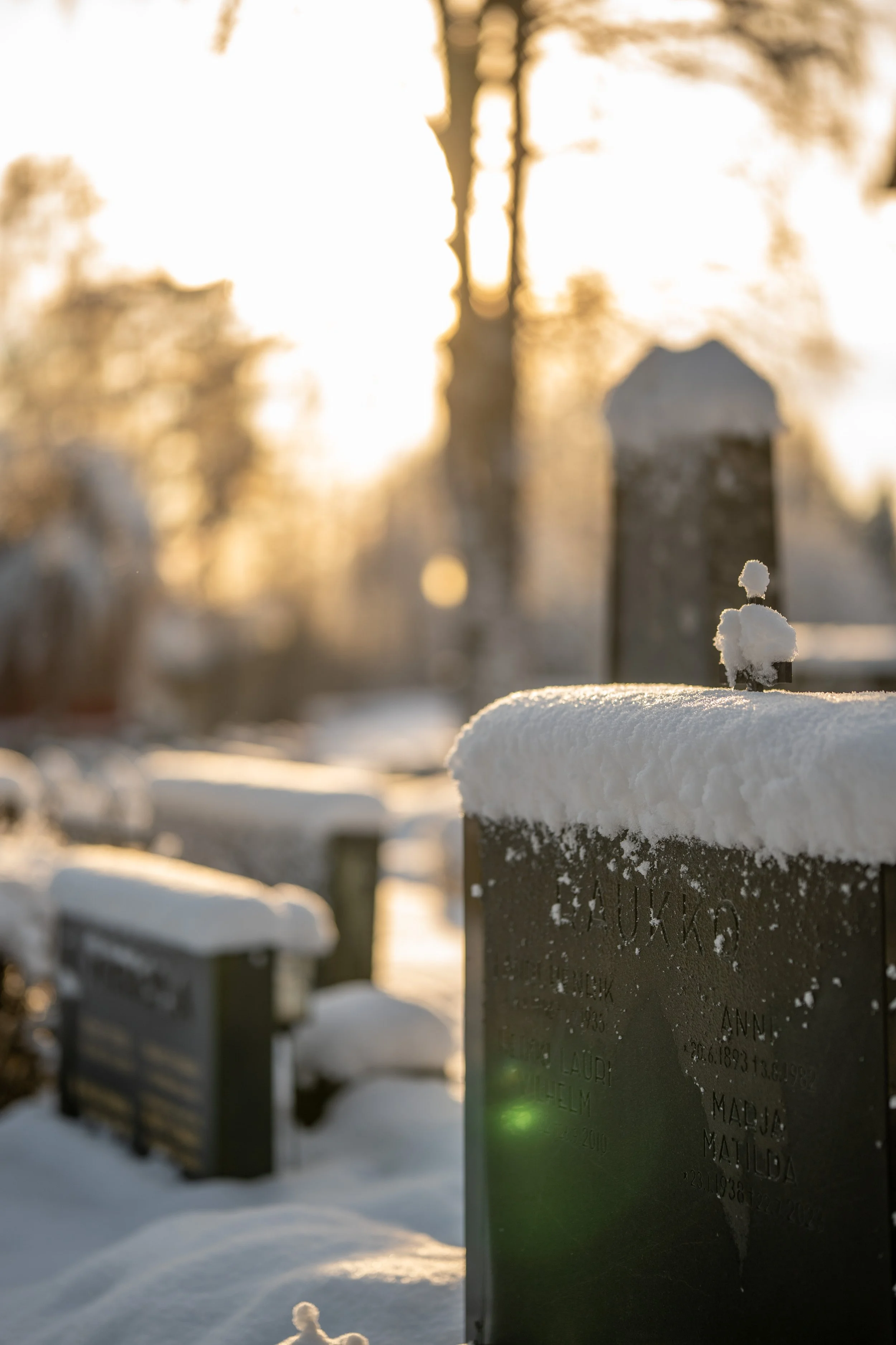 Snow-covered gravestones in a cemetery at sunset with blurred trees in the background.