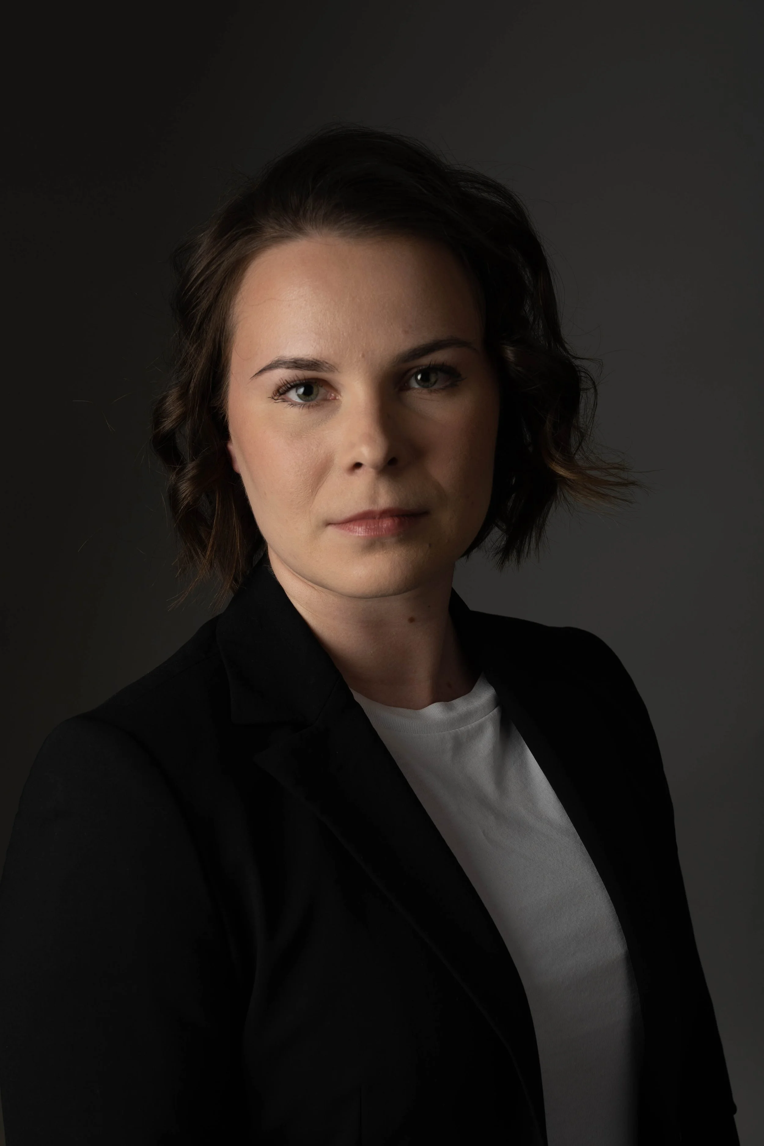 A woman with short, wavy brown hair, wearing a black blazer and a white t-shirt, looking directly at the camera against a dark background.