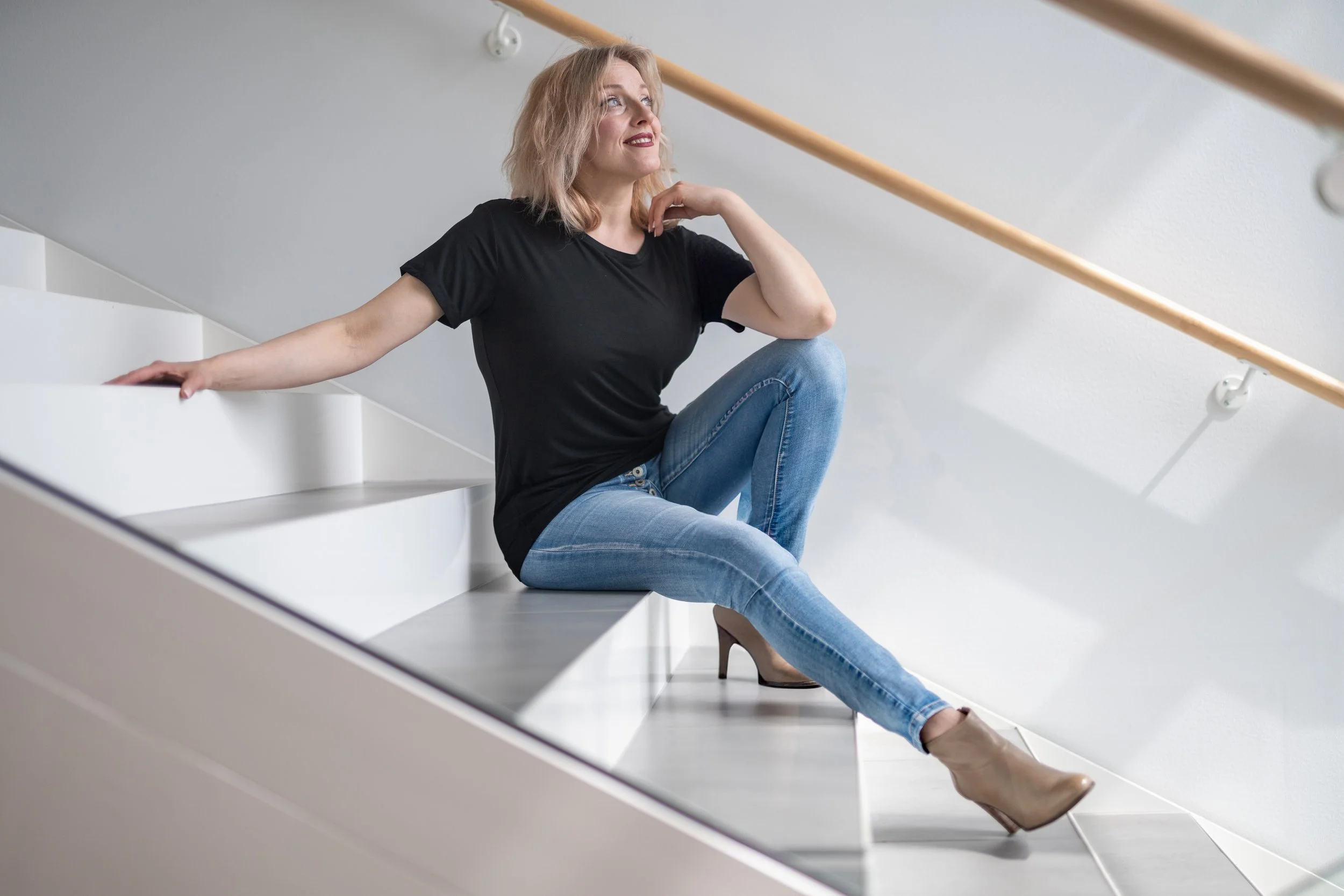 A woman with blonde hair wearing a black t-shirt, light blue jeans, and tan high heels sitting on white stairs with a wooden handrail.
