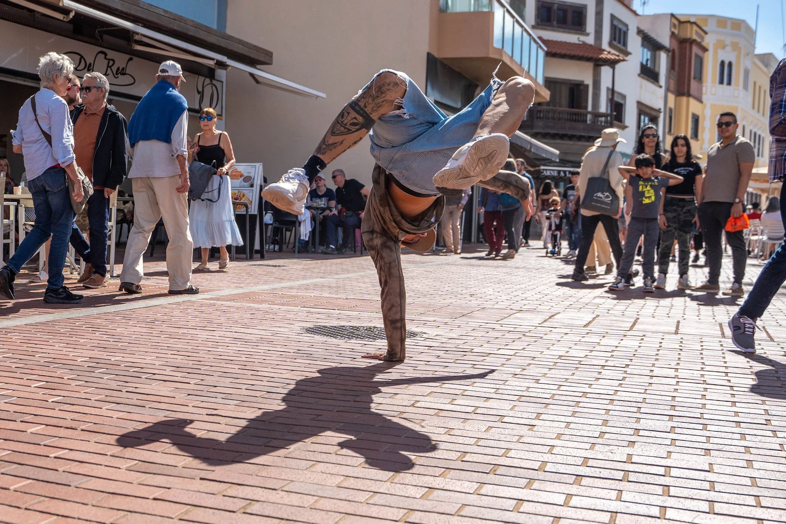 A street performer doing a breakdance move on a brick sidewalk, surrounded by pedestrians and onlookers.