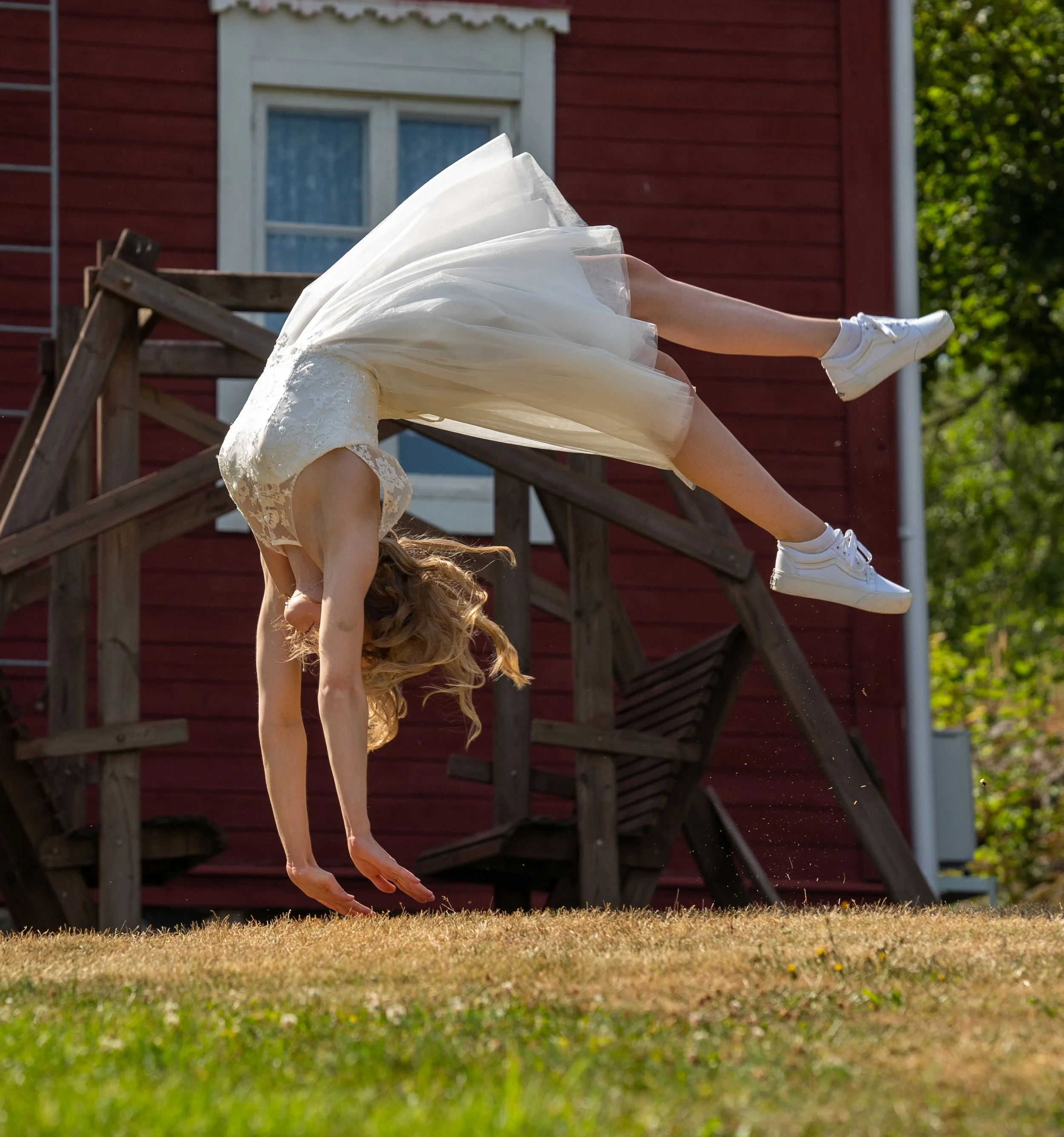 A woman in a white dress and white sneakers is performing a backflip outdoors on grass, with a red wooden structure and green trees in the background.