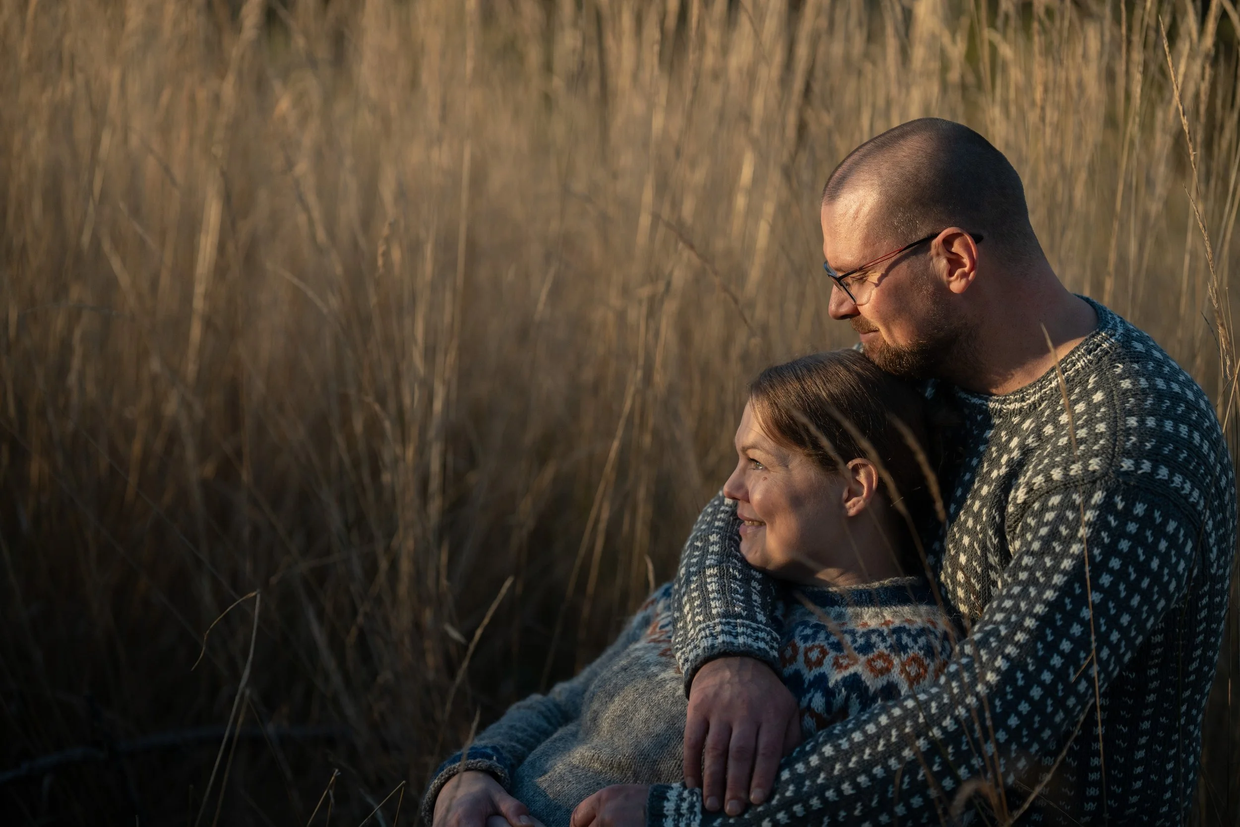 A man with glasses and a woman with red hair smiling and hugging each other in a field of tall grass during sunset.
