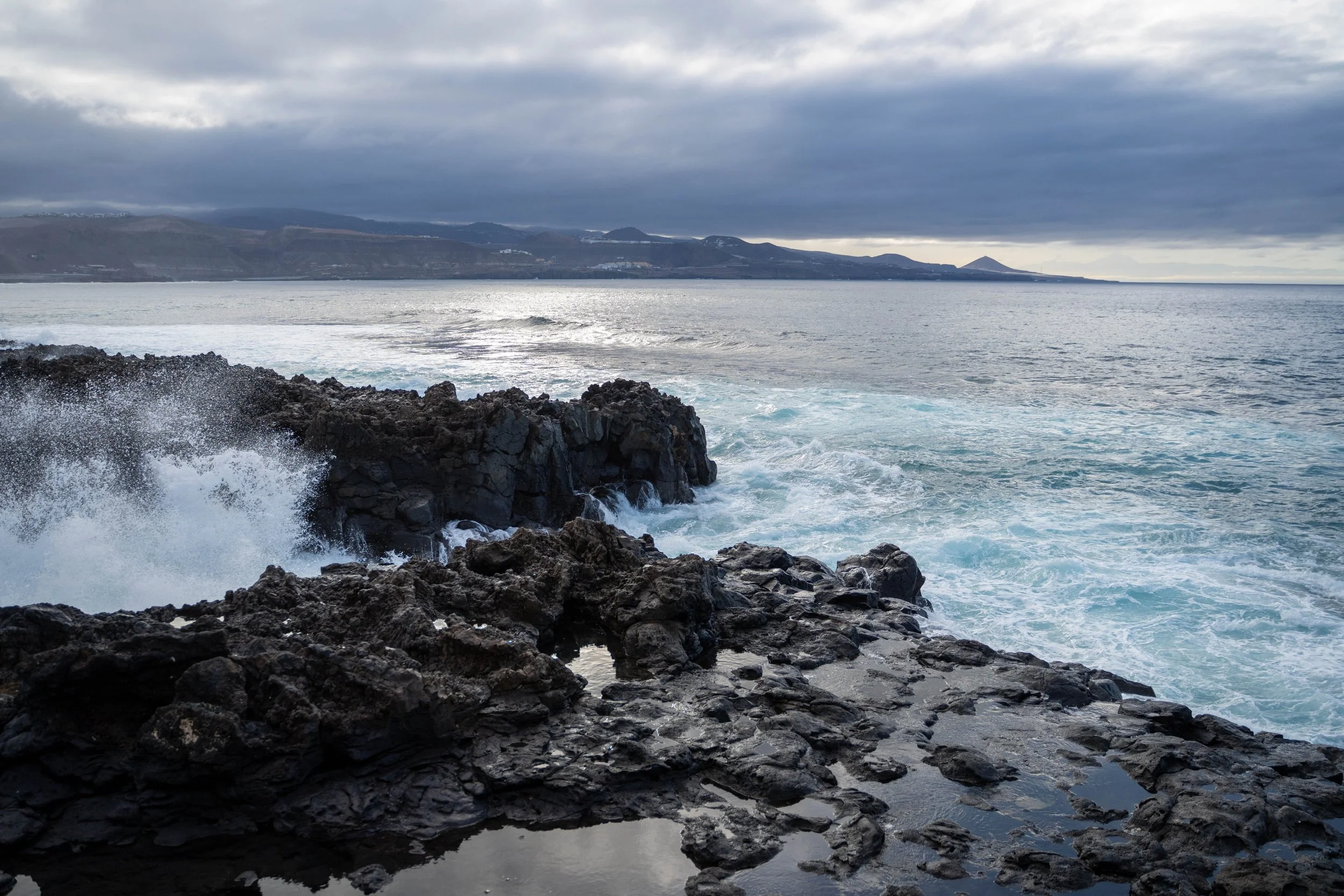 View of the ocean with waves crashing against dark rocky shoreline under cloudy sky, with distant hills in background.