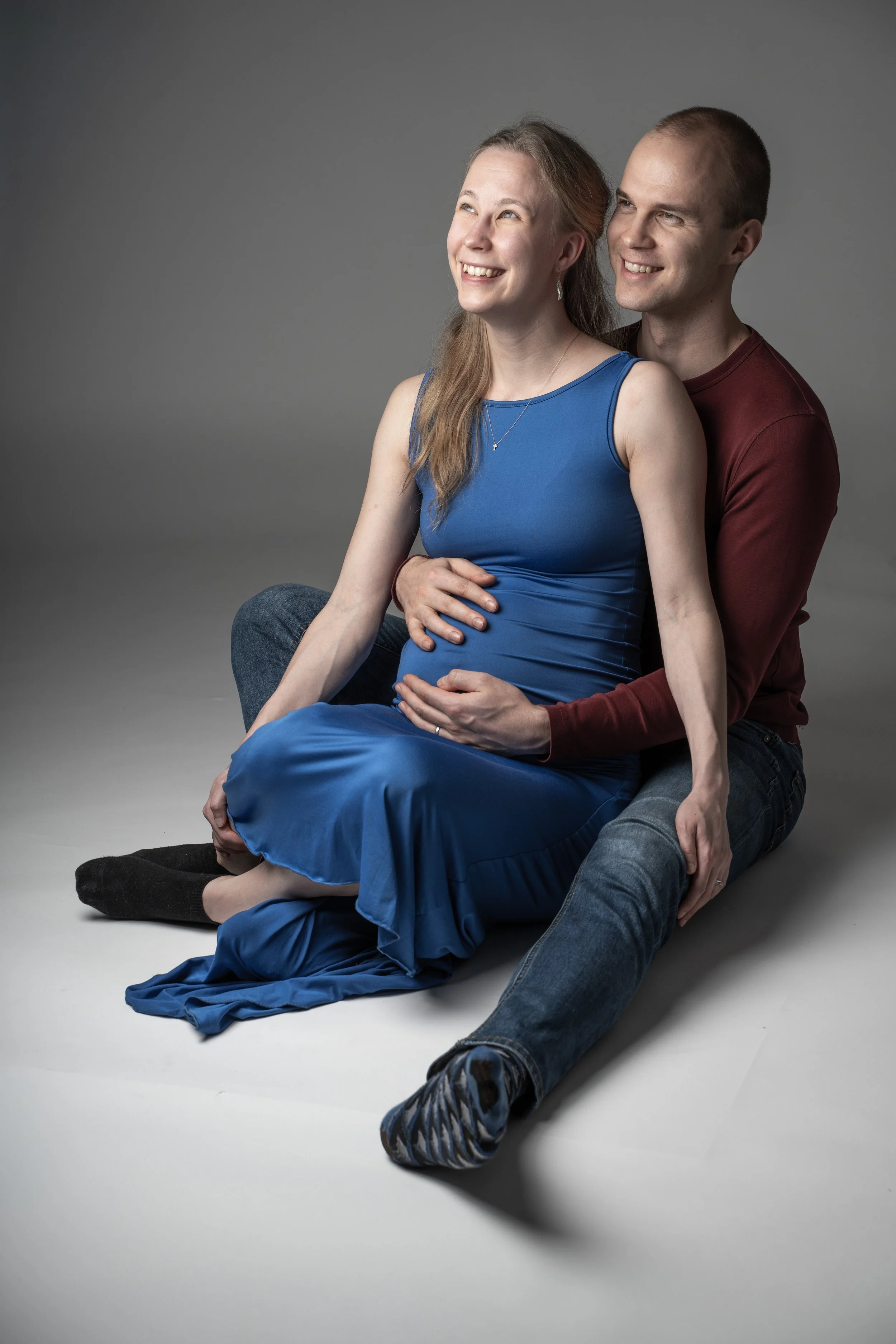 A pregnant woman sitting on the floor, smiling, with her partner behind her, both appearing happy and embracing each other against a gray background.