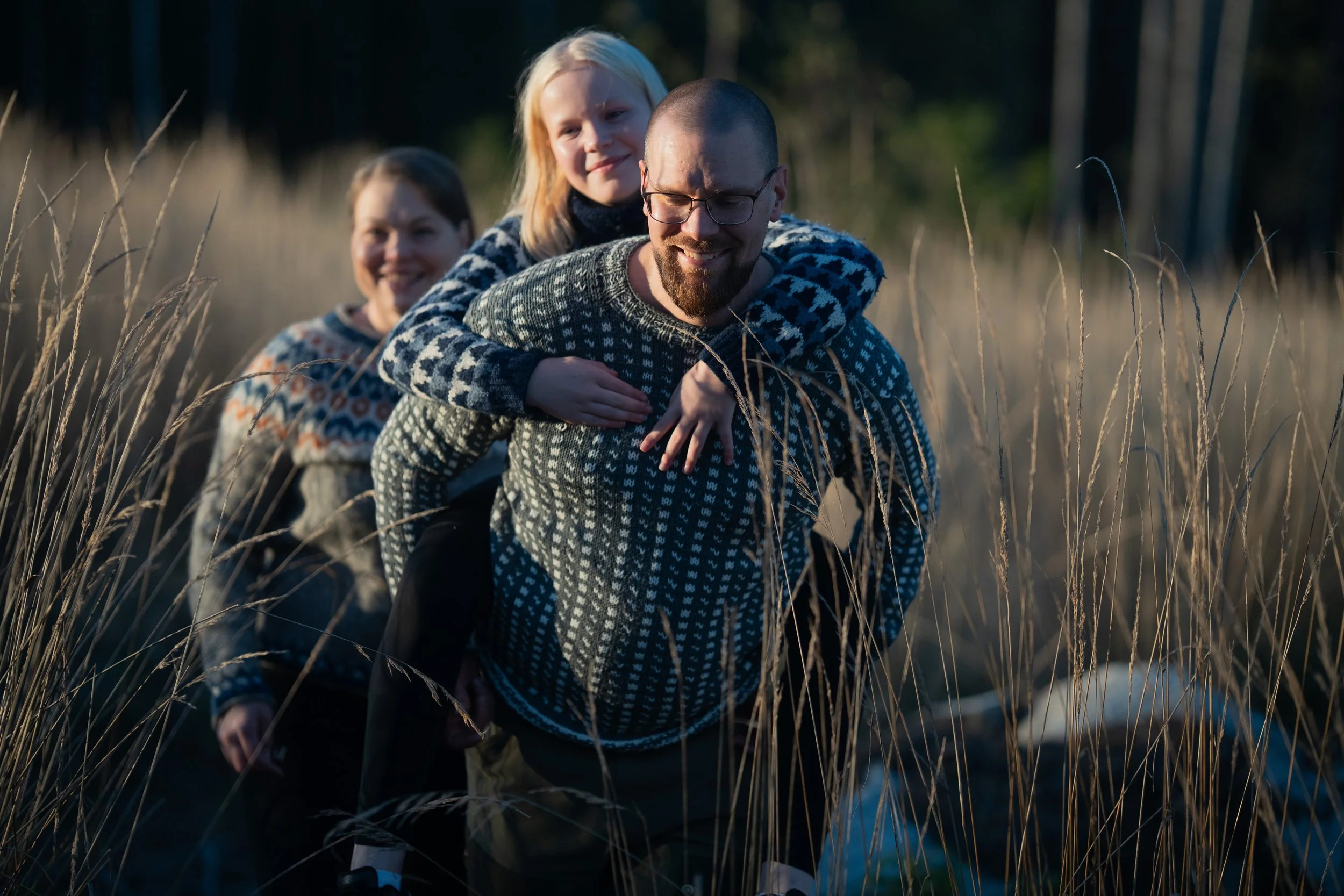 A man giving a piggyback ride to a girl, followed by two women, in a field of tall grass during sunset.