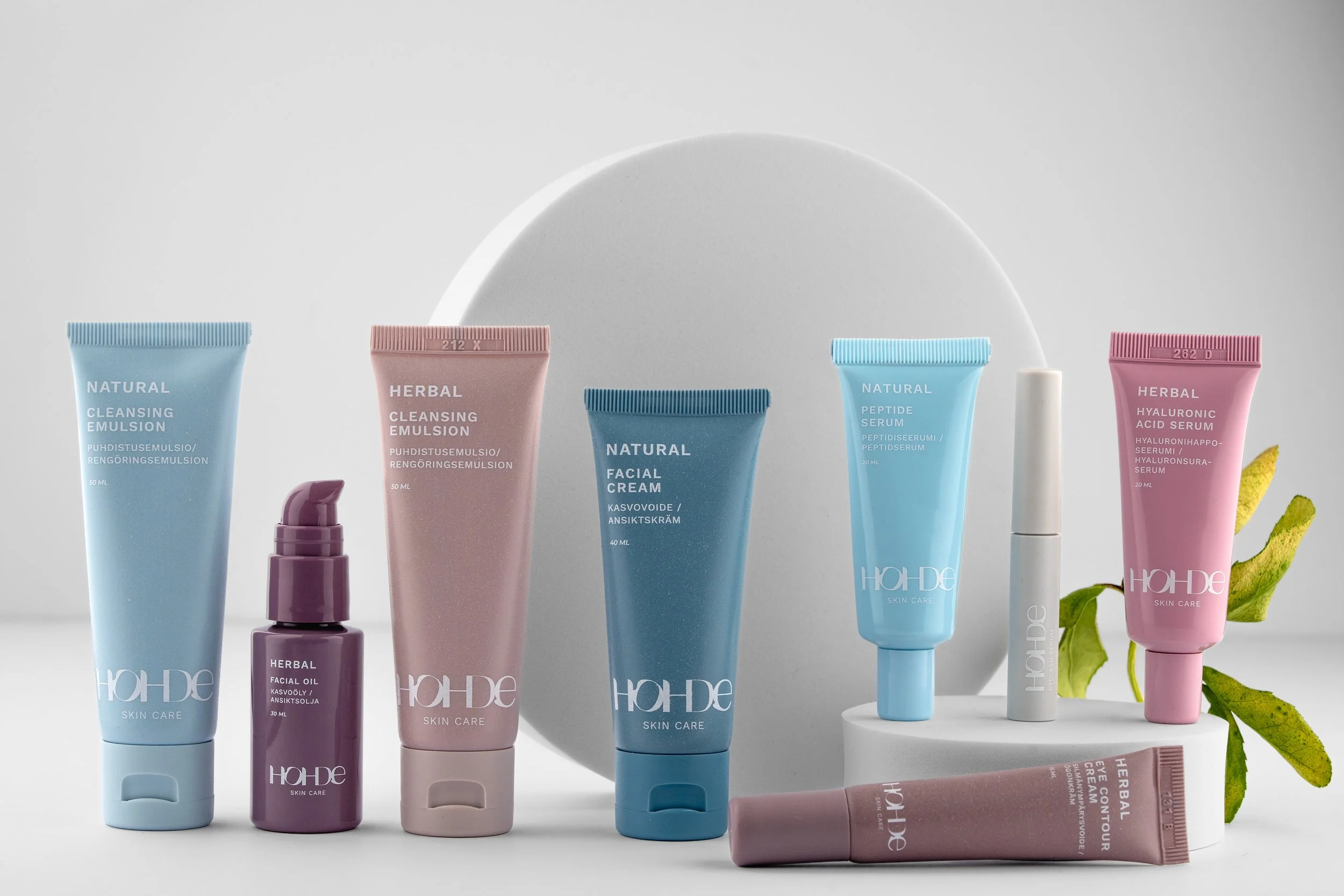 A collection of skincare products, including cleansers, serums, facial cream, and eye contour cream, arranged on a white surface with a white circular background and a green leaf.