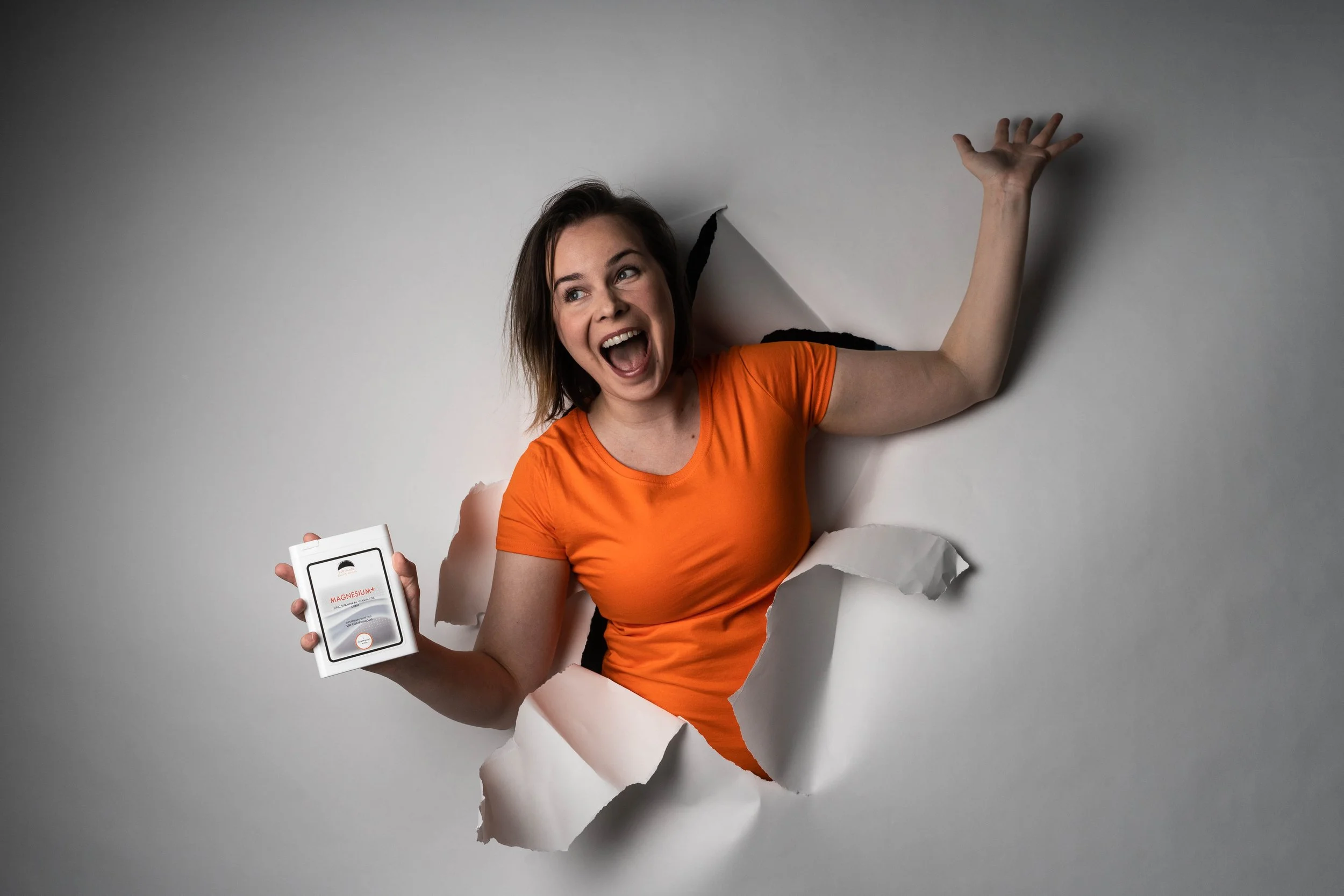 A woman with shoulder-length brown hair, laughing and smiling, wearing an orange t-shirt, coming through a torn piece of white paper wall, holding a bottle labeled 'Magnesium' in her right hand.