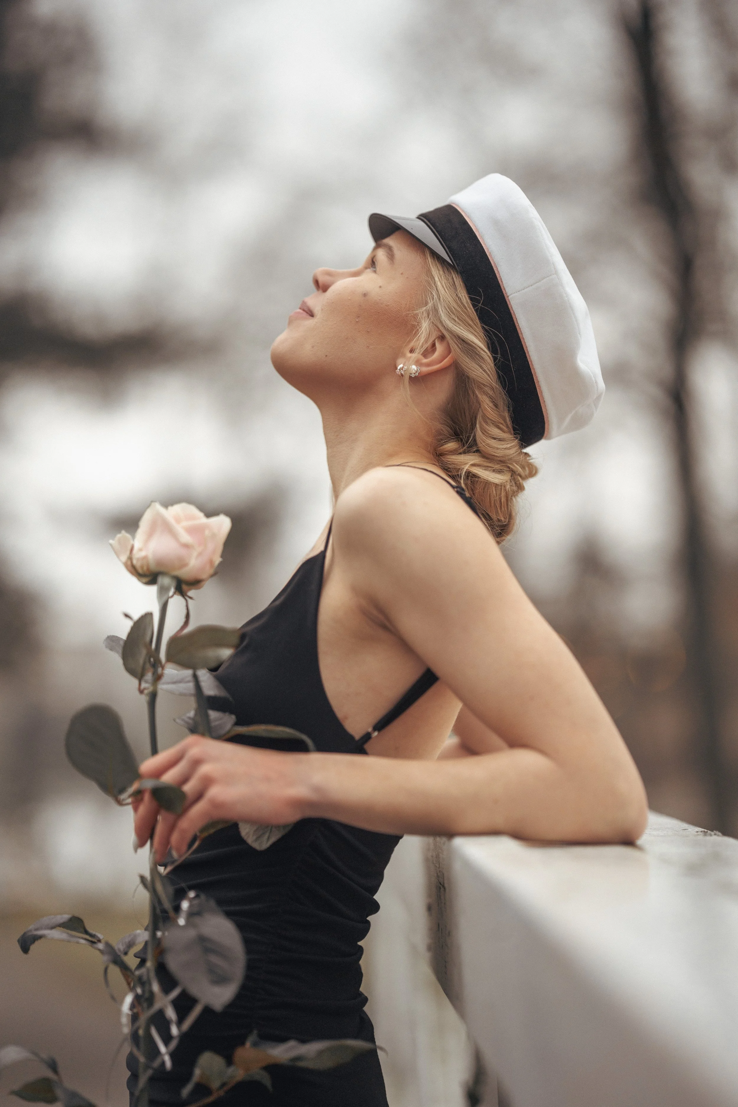 A woman wearing a white and black cap, earrings, and a black sleeveless dress, holding a pink rose, leaning on a white railing outdoors with blurred trees in the background.