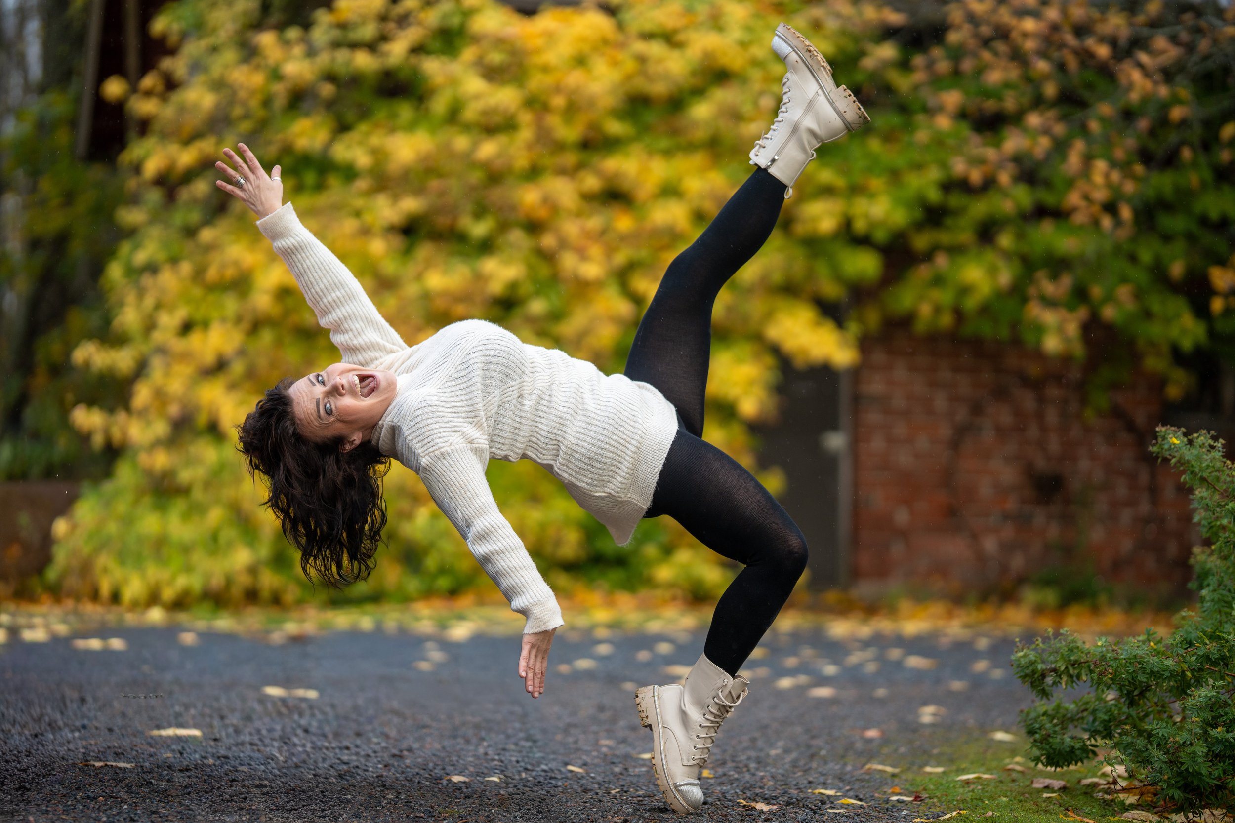 Woman with dark curly hair wearing a white sweater, black leggings, and beige boots, jumping outdoors on a paved path with autumn-colored trees in the background.