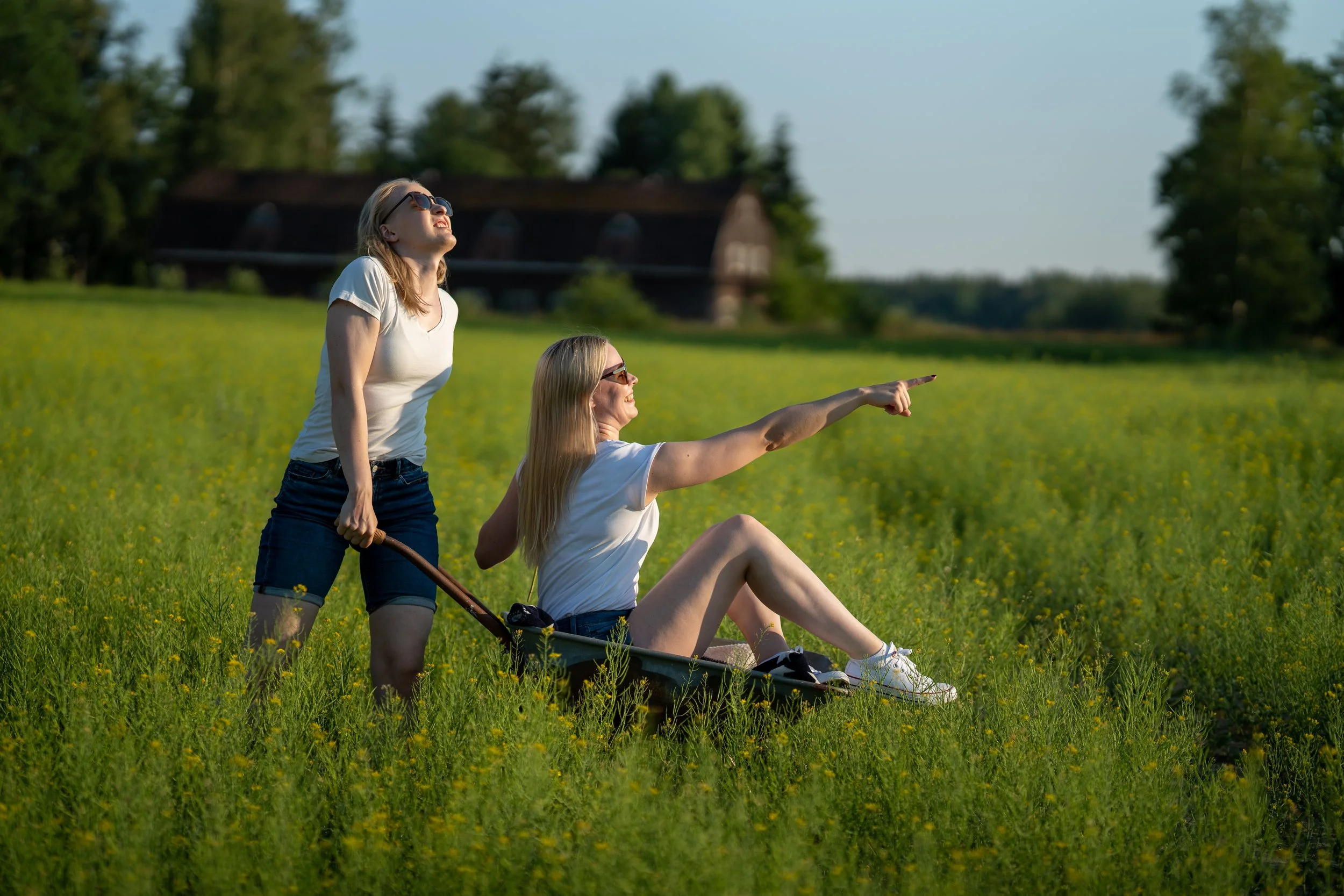 Two young women enjoying a sunny day in a field, one sitting in a wheelbarrow and pointing ahead, the other standing behind holding the wheelbarrow's handle, both wearing casual clothes and sunglasses, with a barn and trees in the background.